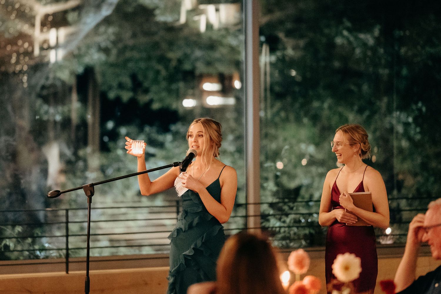 People giving speeches at an evening outdoor reception with string lights and microphone setup.