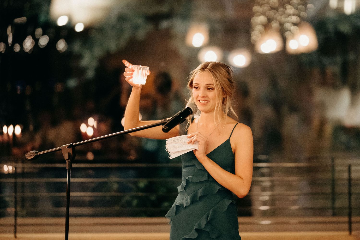 Woman in dark green dress gives speech at evening event holding up glass for toast in front of microphone.