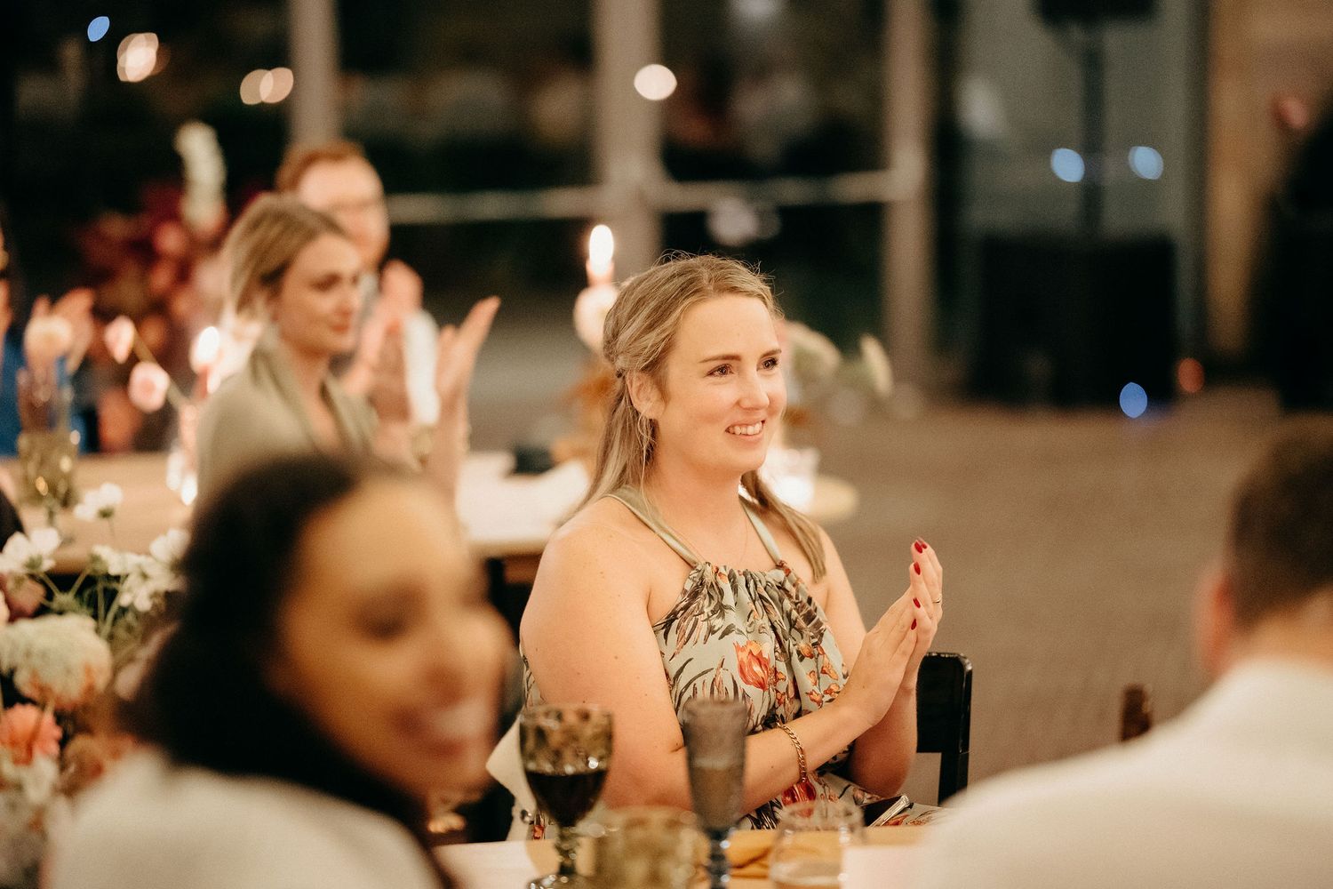 A guest claps enthusiastically at an elegant dinner party with wine glasses and soft lighting in the background.