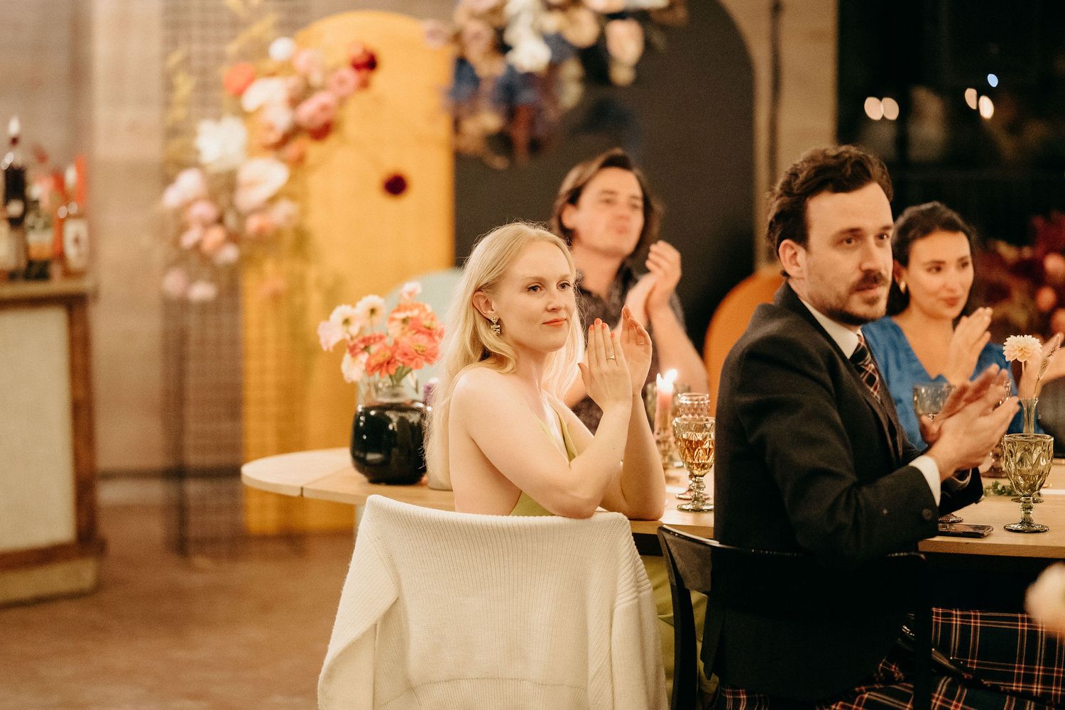 Wedding guests applaud during an elegant evening reception with warm lighting and floral decorations.