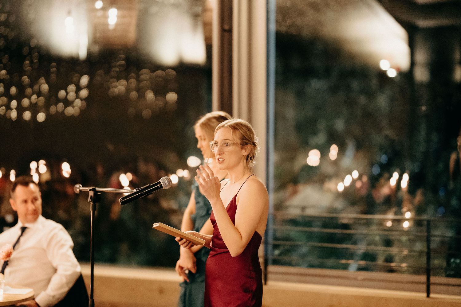 Someone speaking emotionally into a microphone at an evening wedding reception with twinkling lights.