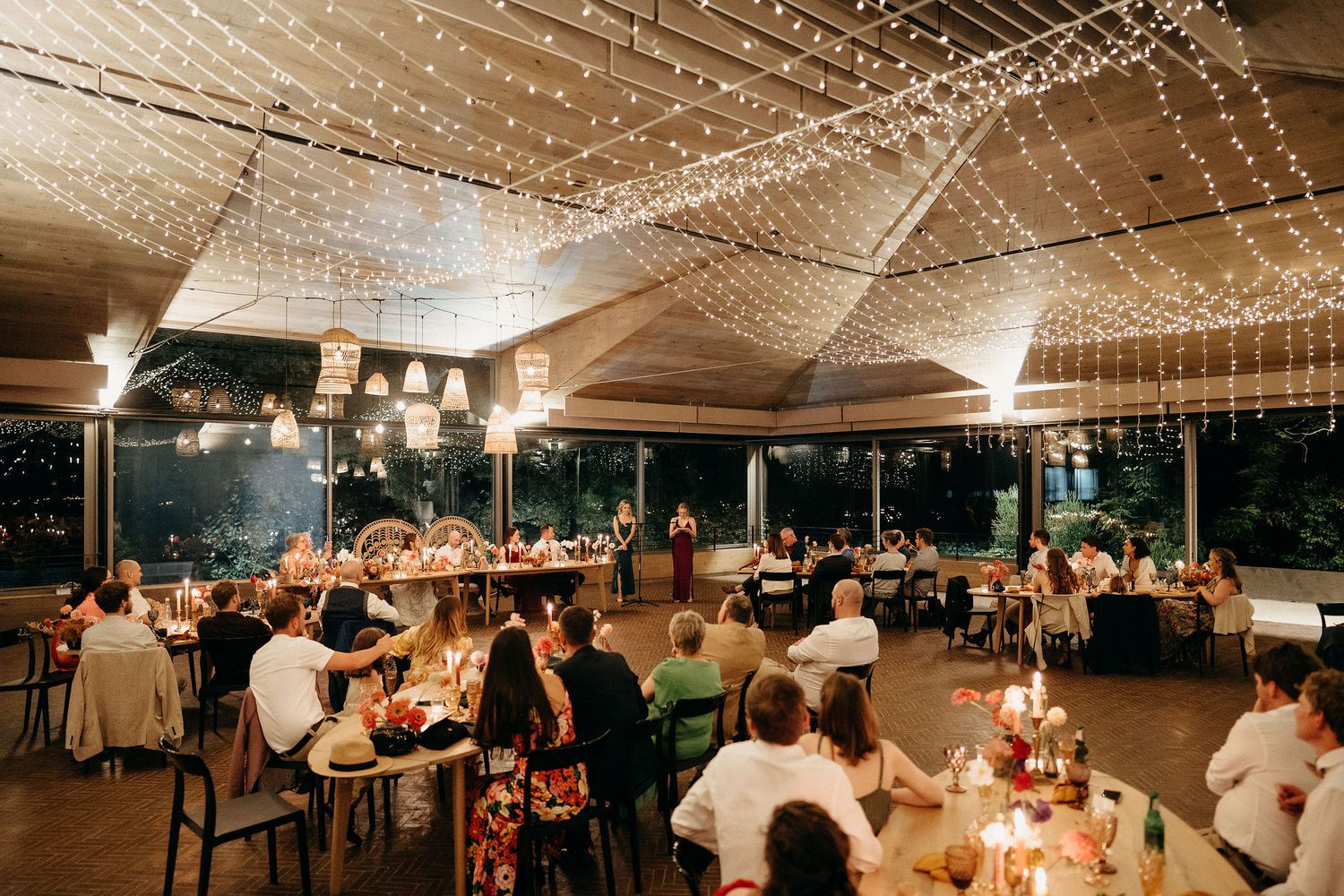 Evening wedding reception under string lights with guests dining at round tables in an elegant covered venue.