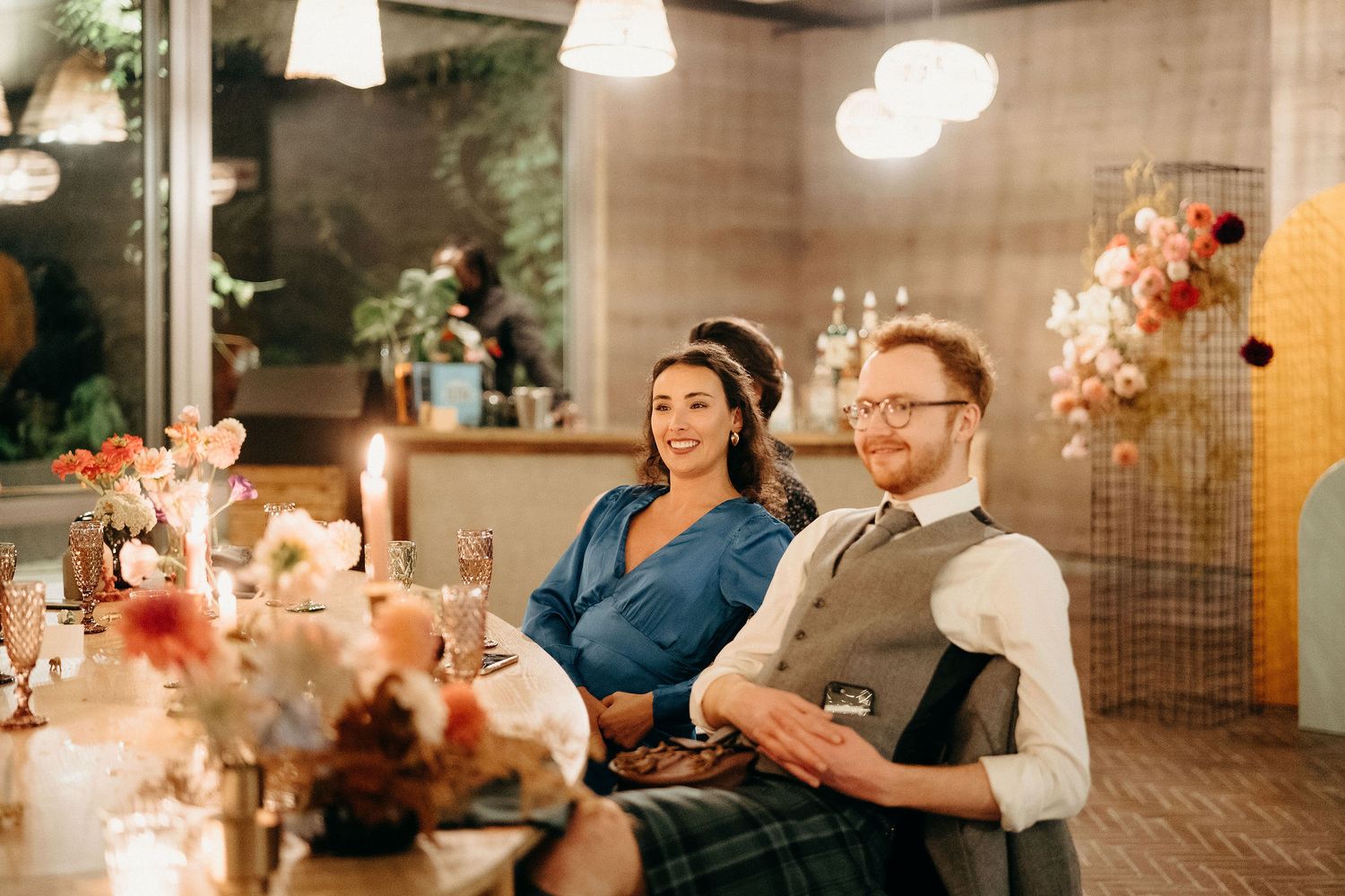 Two guests enjoy a warm candlelit dinner reception with floral arrangements and soft lighting in an intimate restaurant setting.