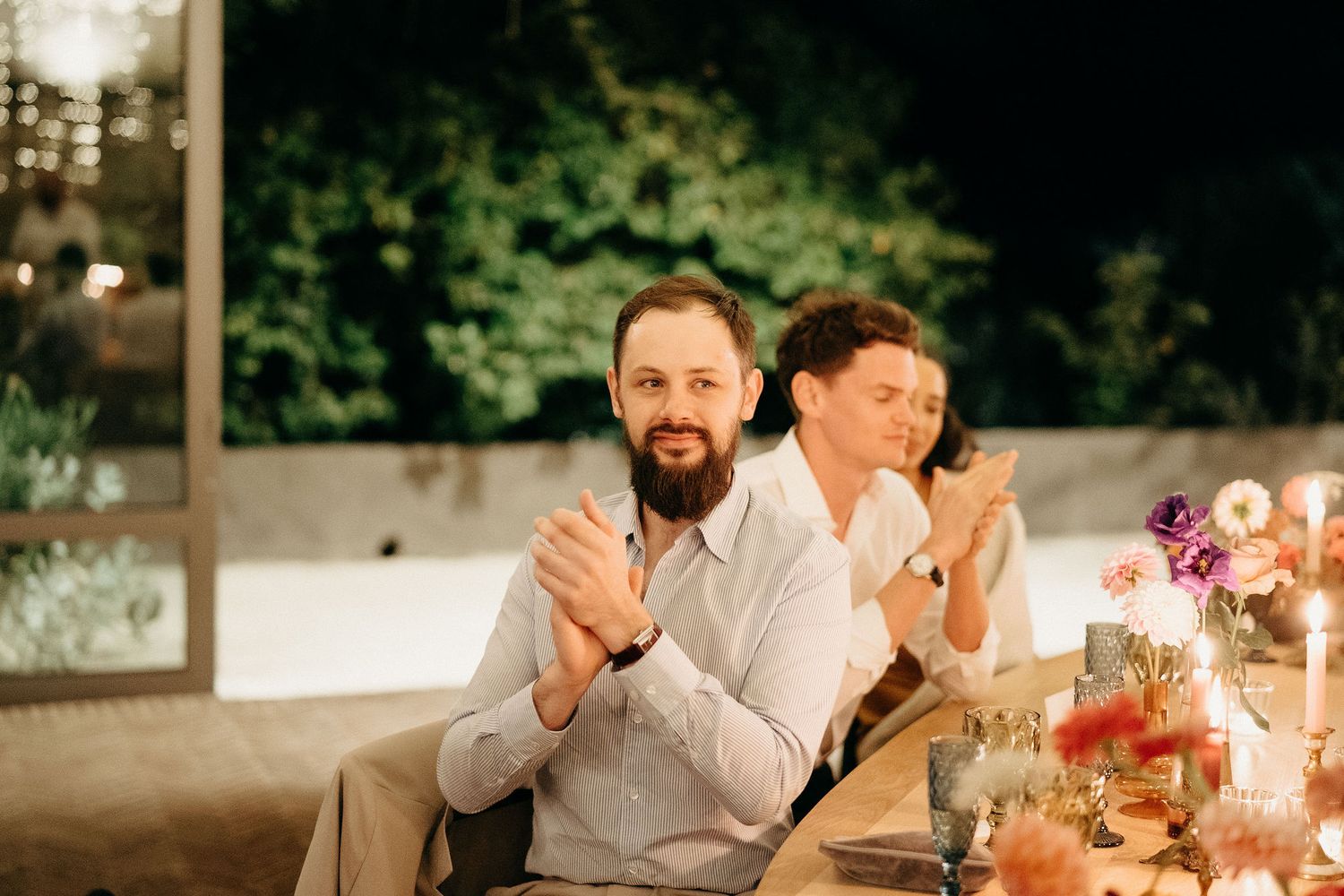Laughter and applause during a festive dinner reception with candlelit table setting in an outdoor venue.