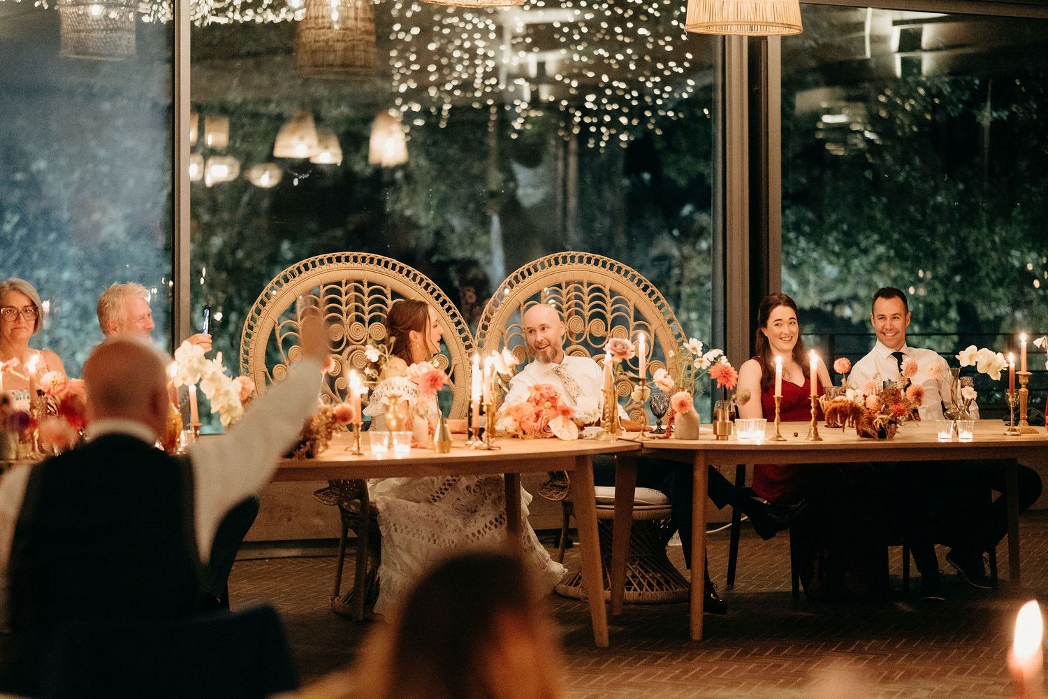 Wedding reception guests seated at a long table under arched decorations and string lights during an evening celebration.