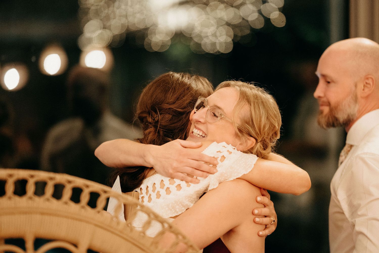 Two people embrace in an emotional hug at an evening celebration while another person looks on in the background.