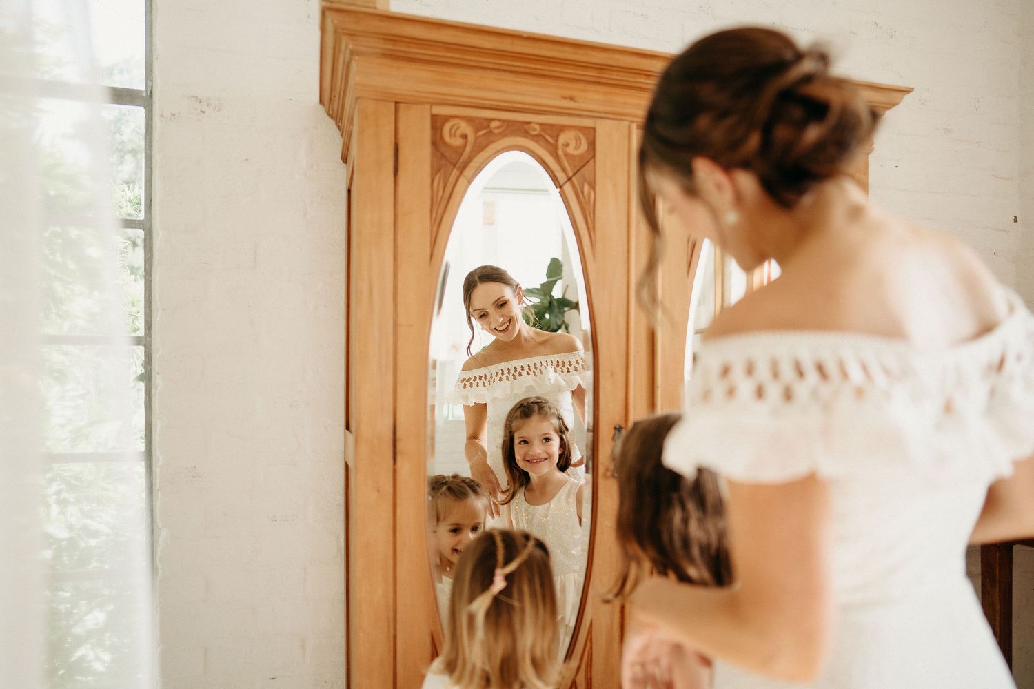 A bride in a white lace dress looks at her reflection in an oval mirror mounted on a wooden wardrobe.