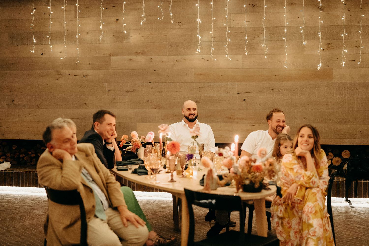 A candid group dinner scene under warm string lights at a rustic wood-paneled restaurant.