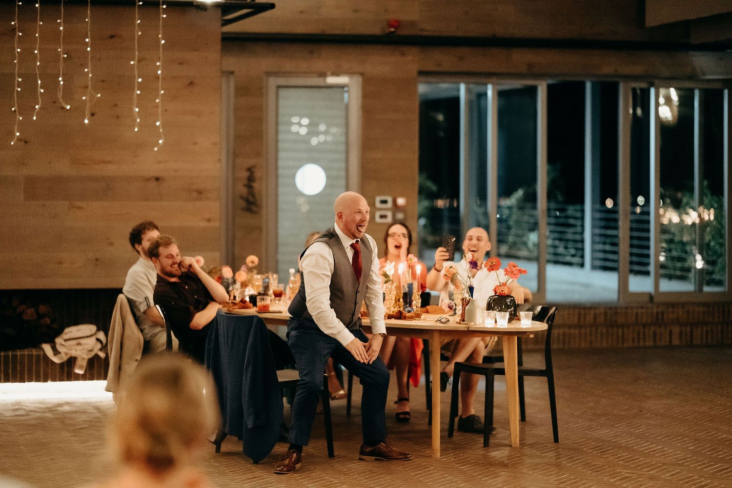 Wedding reception guests enjoy candlelit dinner at long wooden table in rustic indoor venue with string lights.