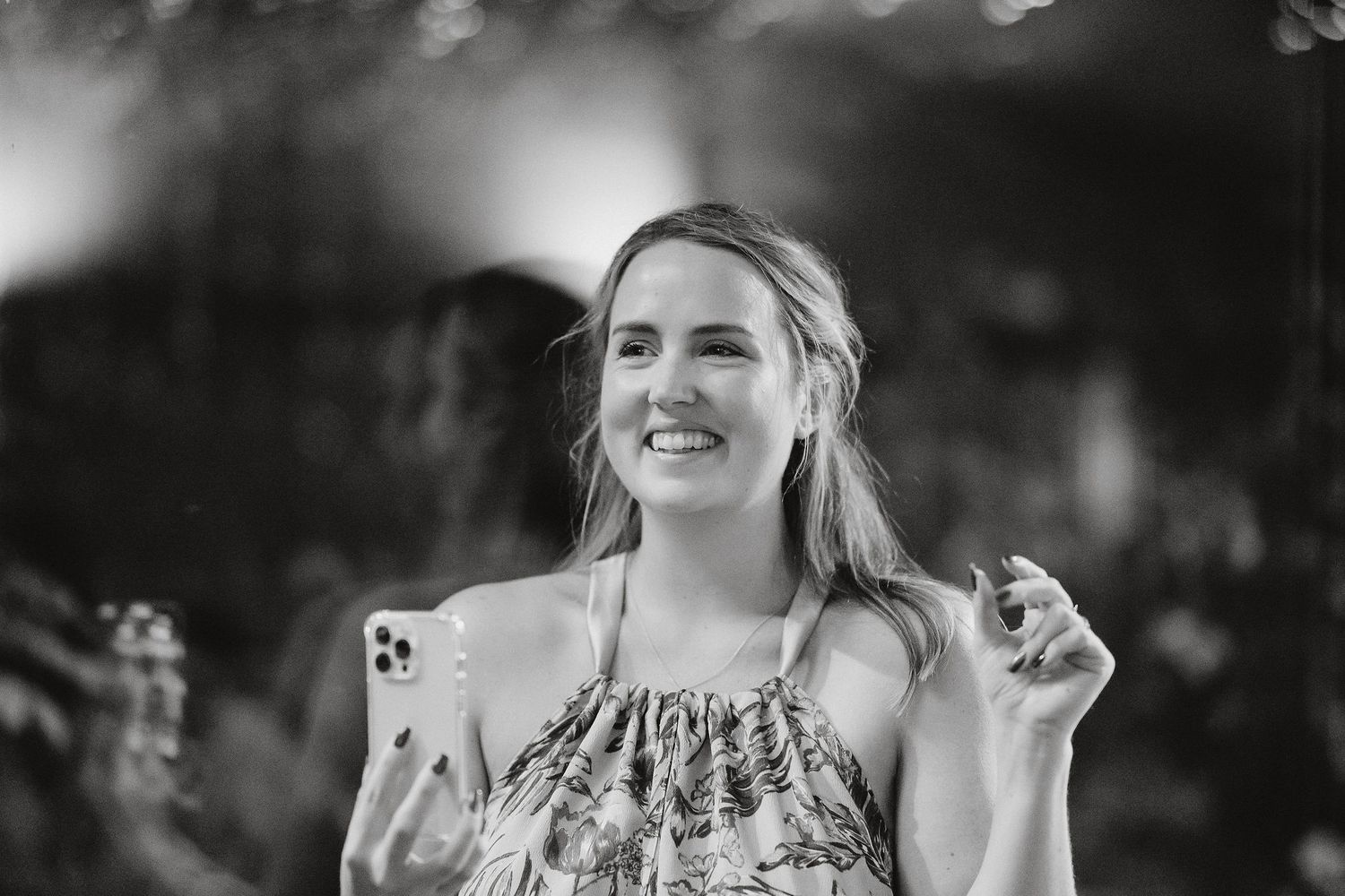 A candid black and white portrait of someone smiling while holding an iPhone in an outdoor setting.