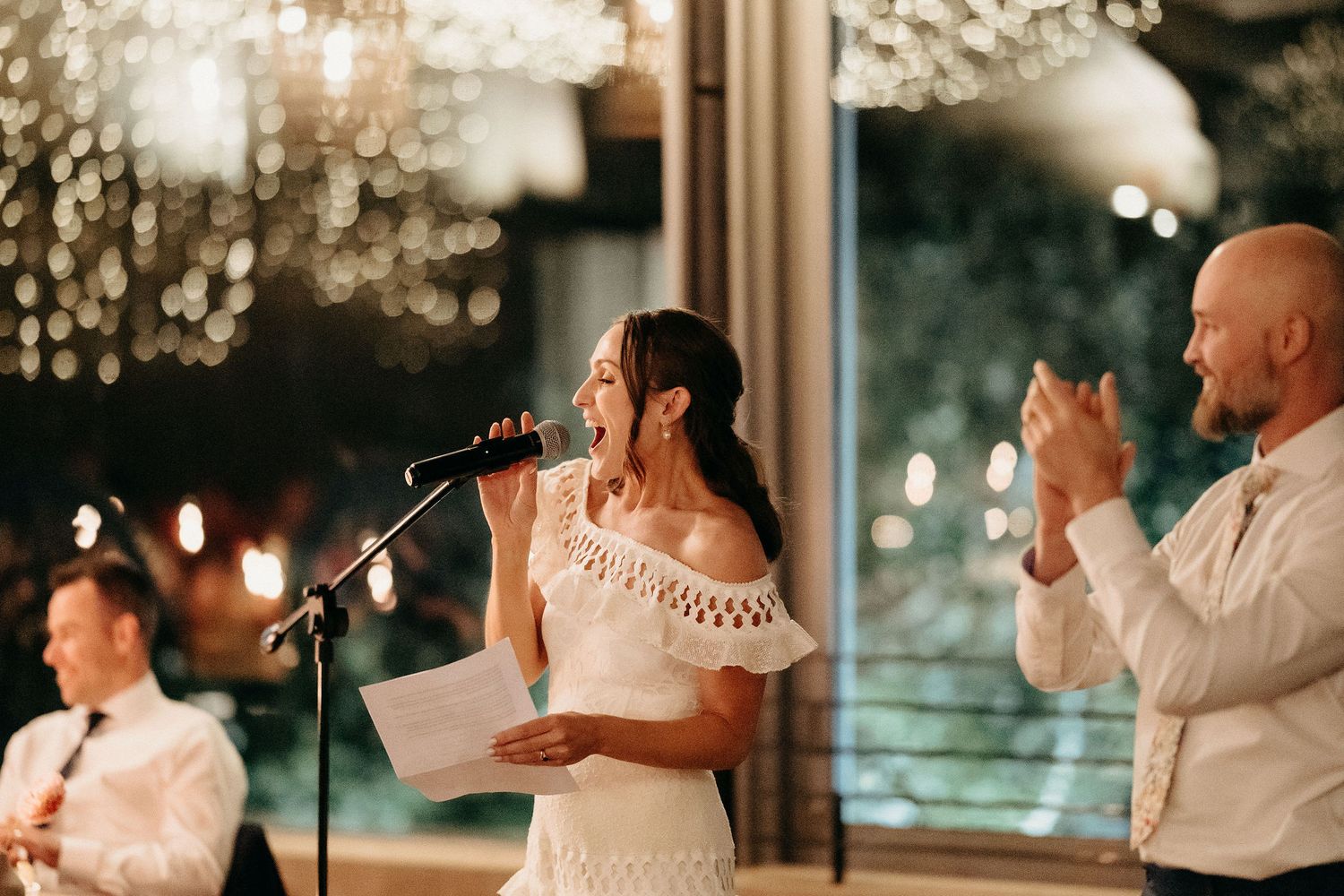 A couple speaks at their wedding reception under a crystal chandelier with soft evening lighting and elegant decor.