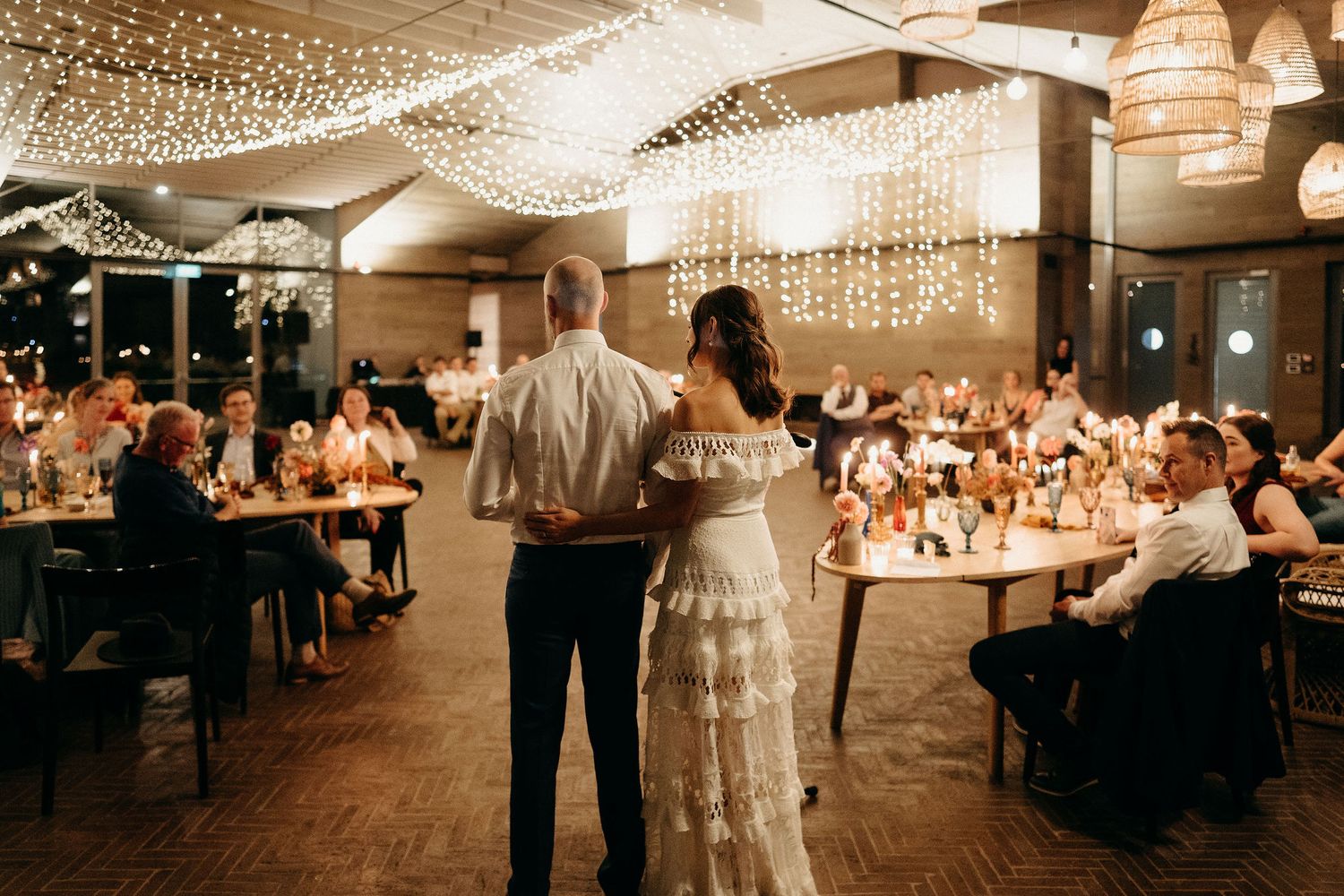 Elegant wedding reception venue decorated with twinkling string lights and lanterns as guests dine at round tables.