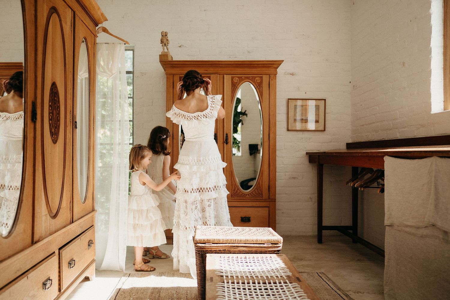A parent and child in white lace dresses look through an antique wooden wardrobe with oval mirror in a rustic room.