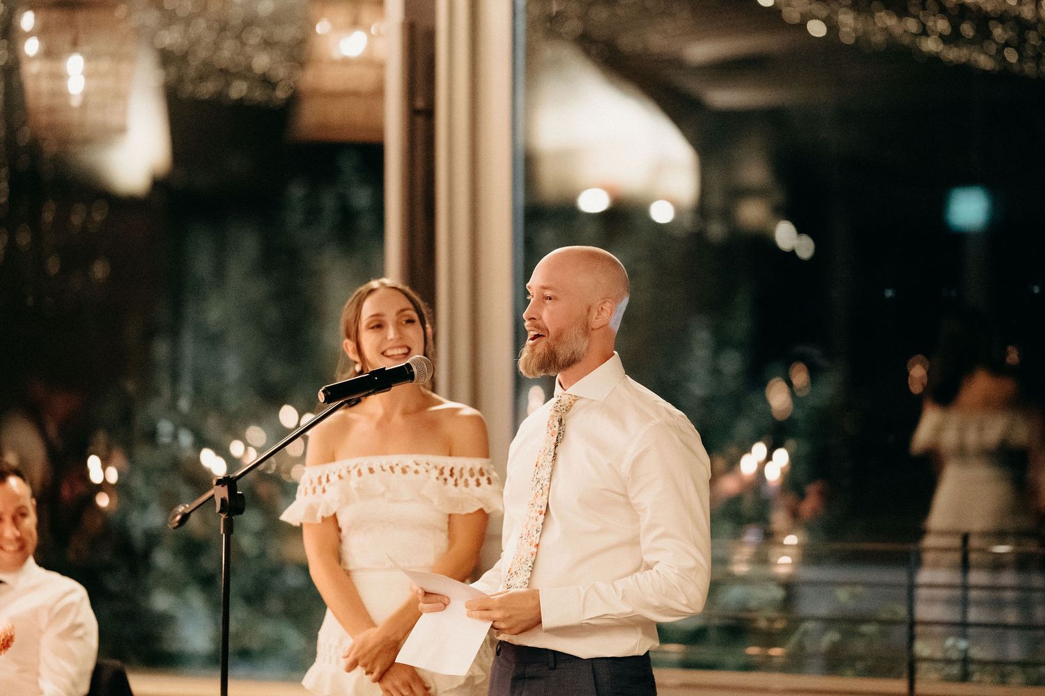 A couple speaks into microphones during an evening reception in an elegantly lit indoor venue.