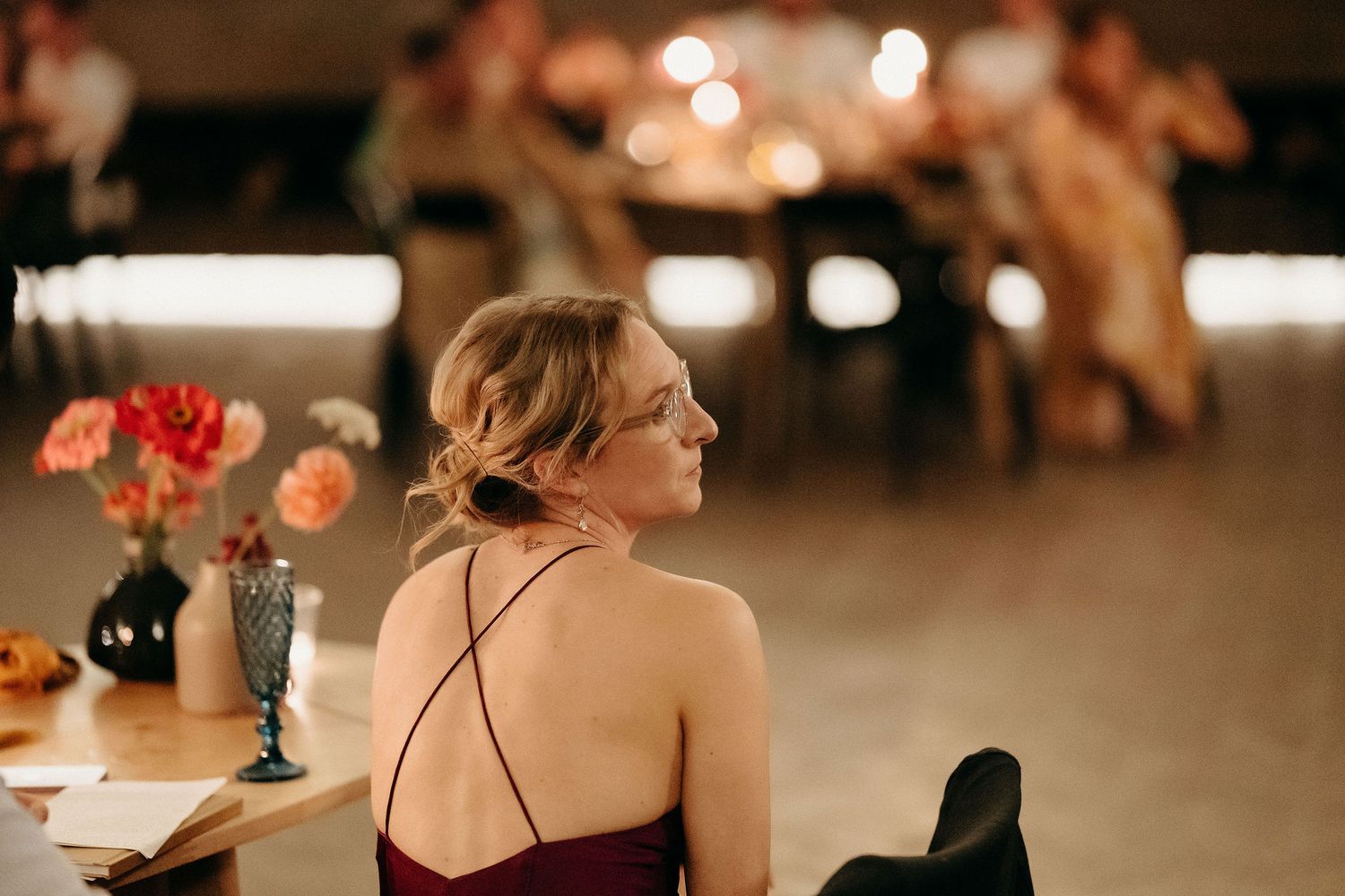 Person in burgundy dress sits at a table with flowers during an evening event with soft bokeh lighting in background.