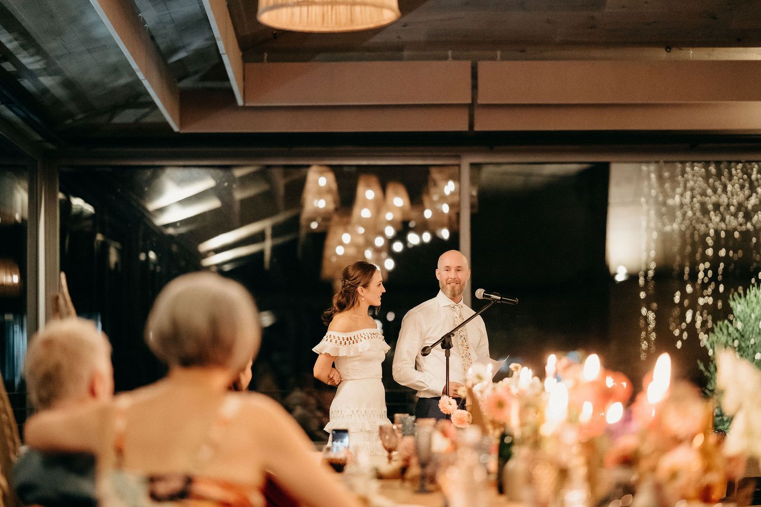 Couple giving heartfelt wedding speeches at an intimate candlelit reception with string lights and romantic ambiance.
