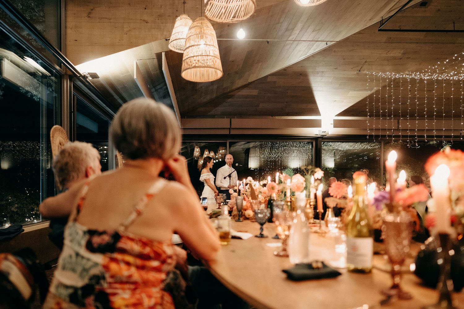 An evening wedding reception with glowing candles and ambient lighting illuminates guests enjoying dinner at a rustic venue.