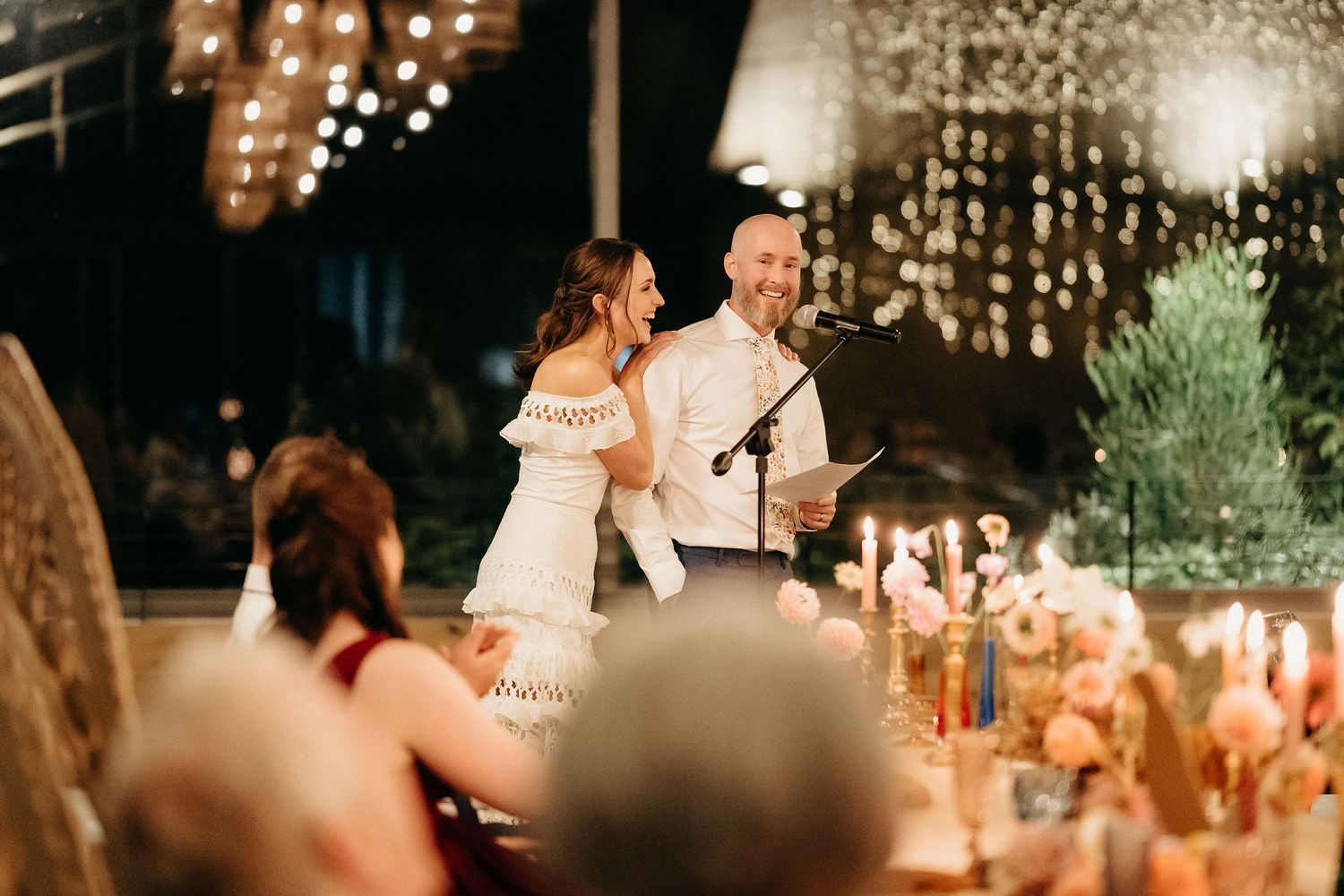 A warm and romantic wedding reception speech illuminated by candlelight and string lights in an elegant venue.