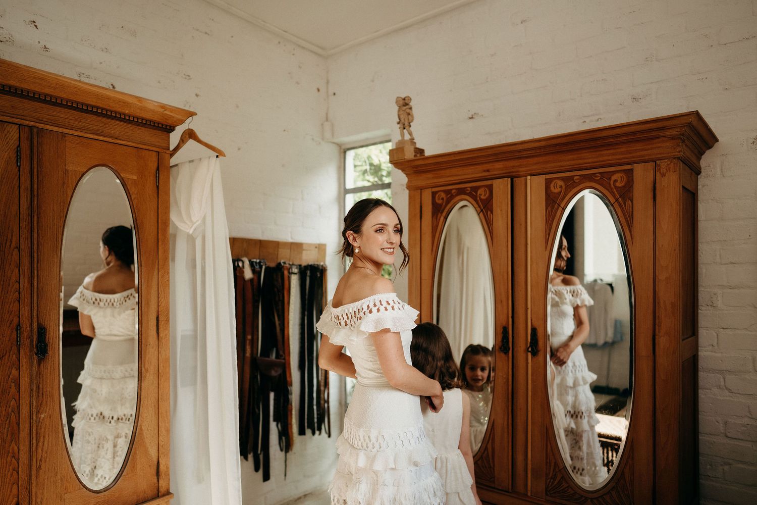 An elegant off-shoulder wedding dress is reflected in vintage wooden wardrobe mirrors in a bright white room.
