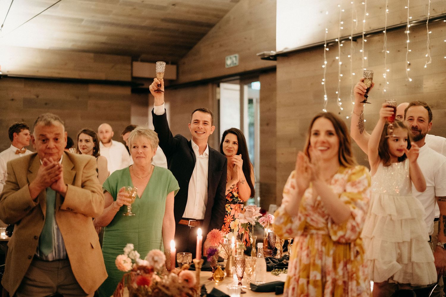 Wedding reception guests celebrate with raised glasses during a toast in a rustic wooden venue with string lights.