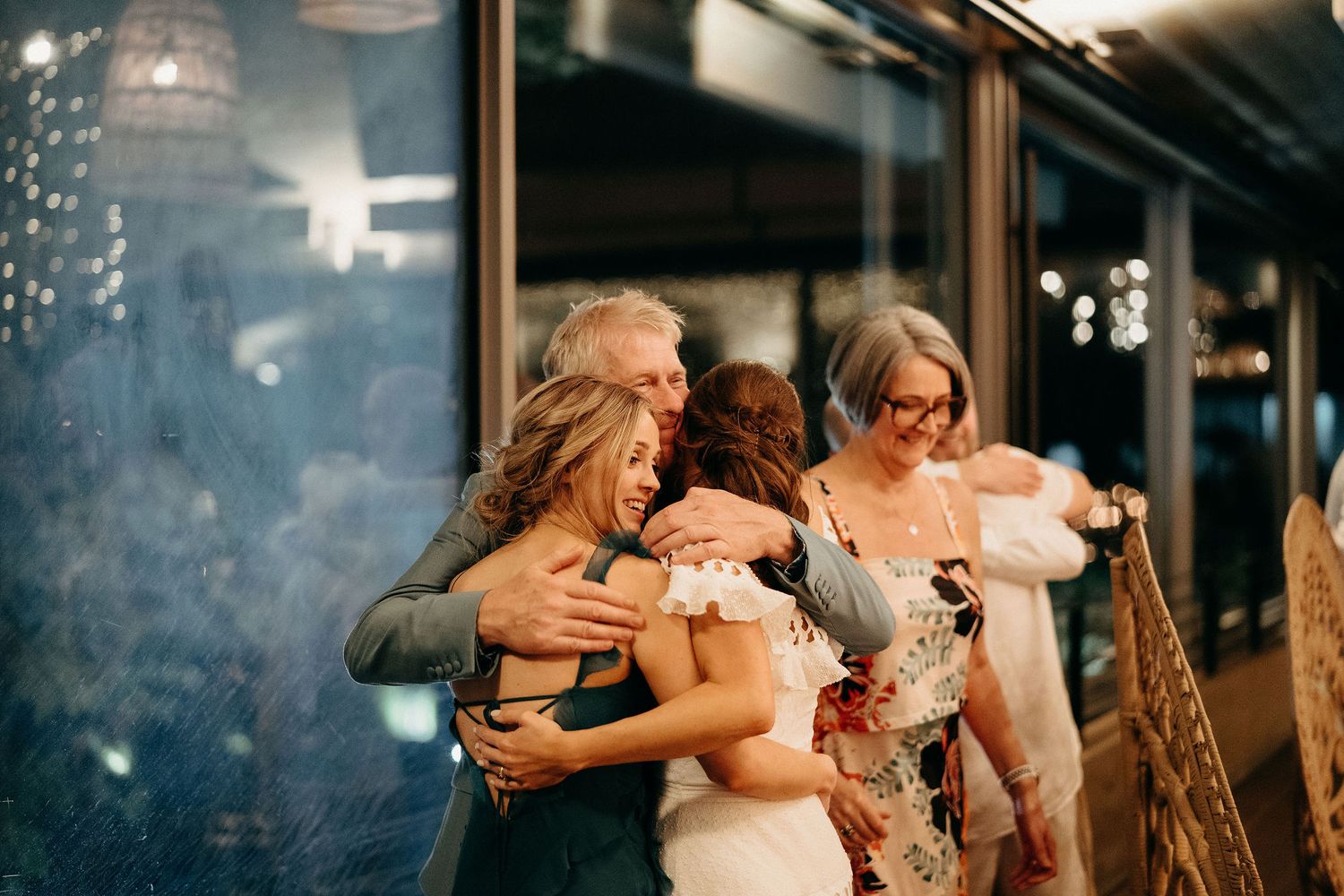 Three people share an emotional embrace during an evening celebration with glass walls and twinkling lights in the background.