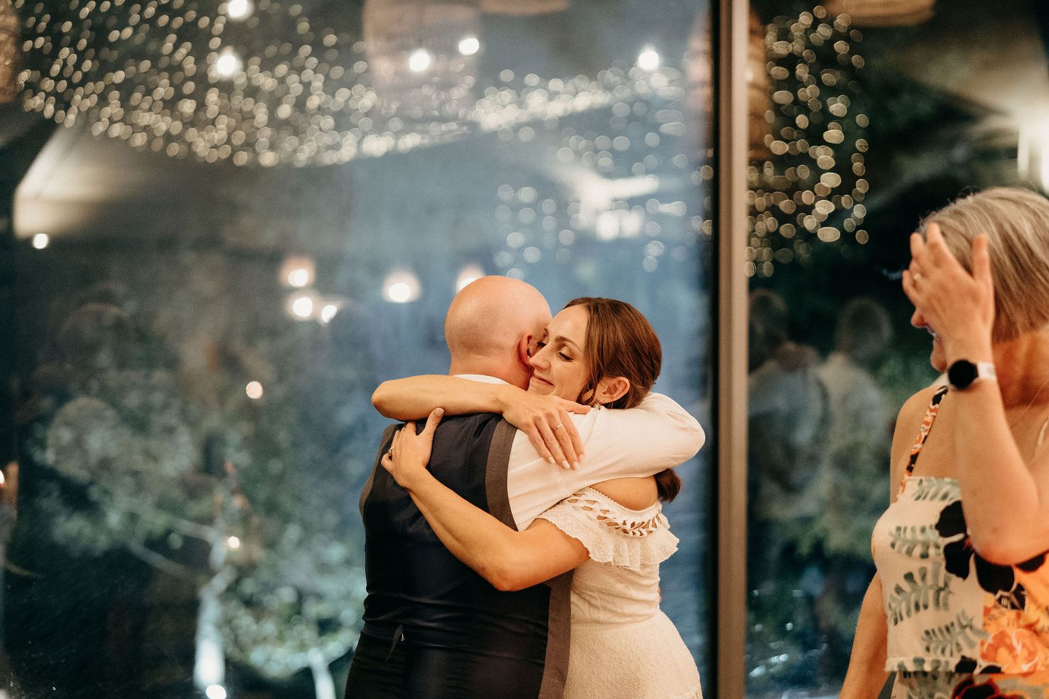 A loving embrace on a dance floor with twinkling lights and guests watching the emotional moment at a wedding reception.