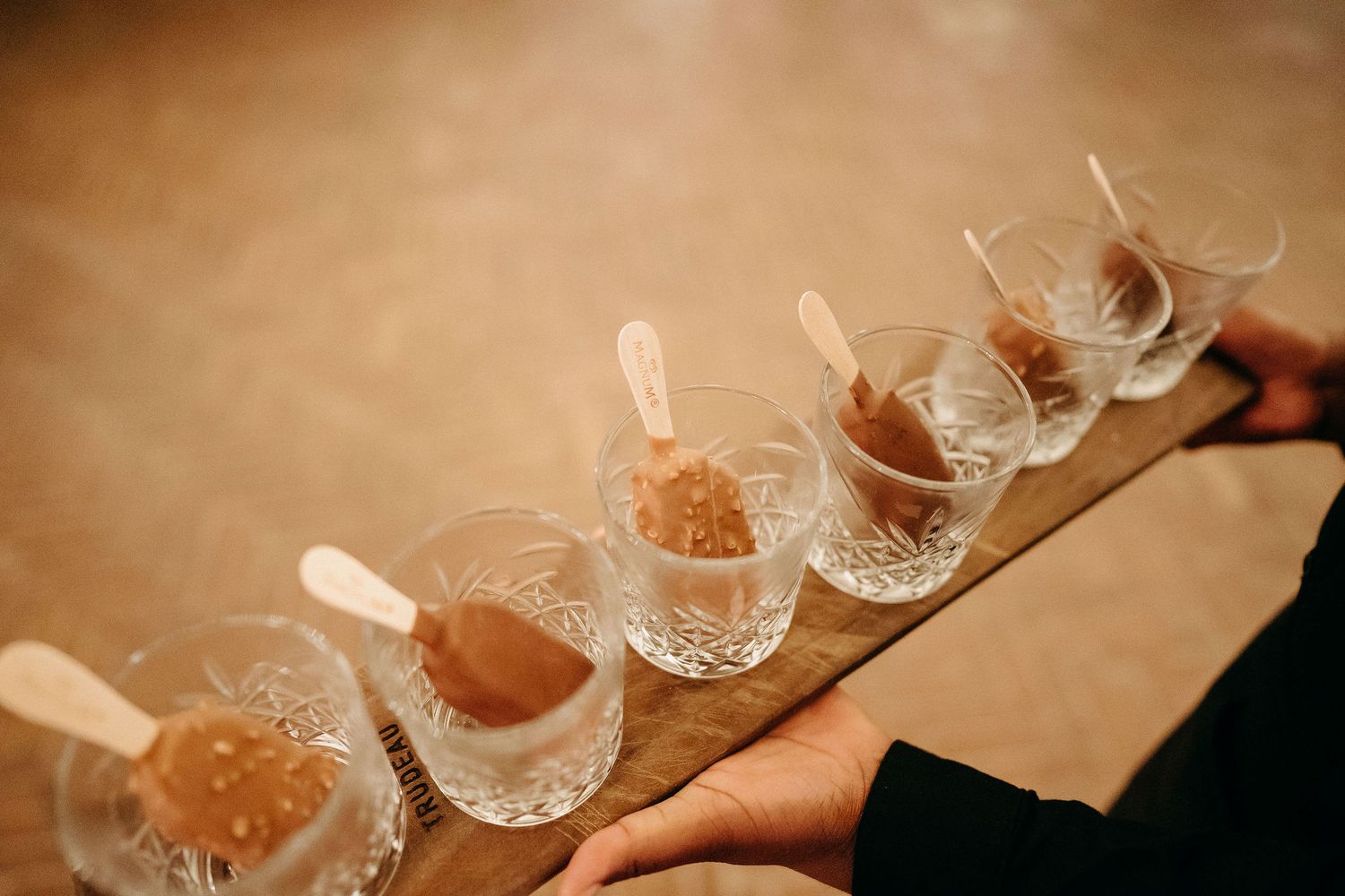 Row of small crystal glasses filled with dessert shots arranged on a wooden serving board being presented.