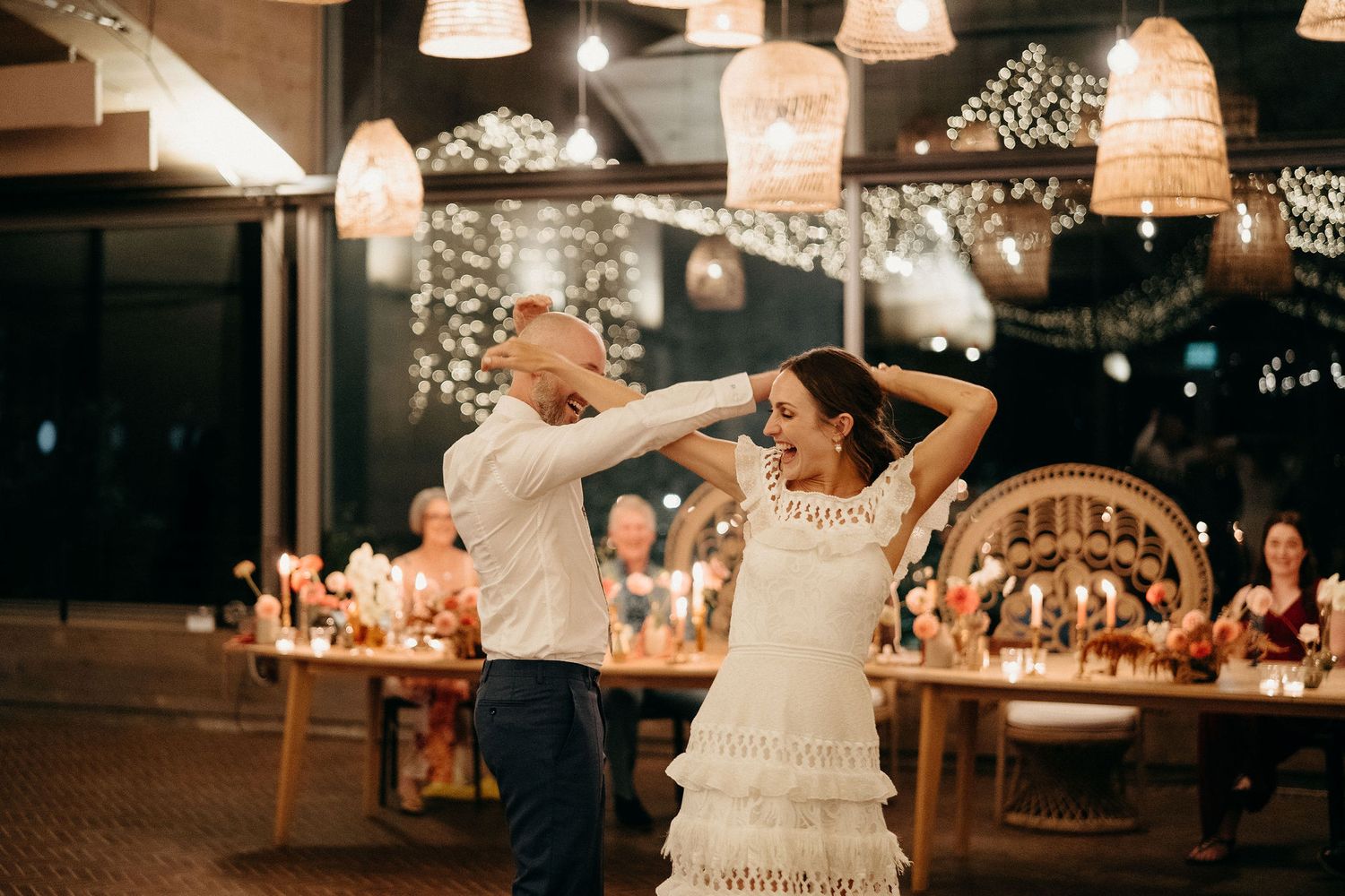 Dancing couple twirls beneath warm string lights and woven lamp shades during a wedding reception celebration.