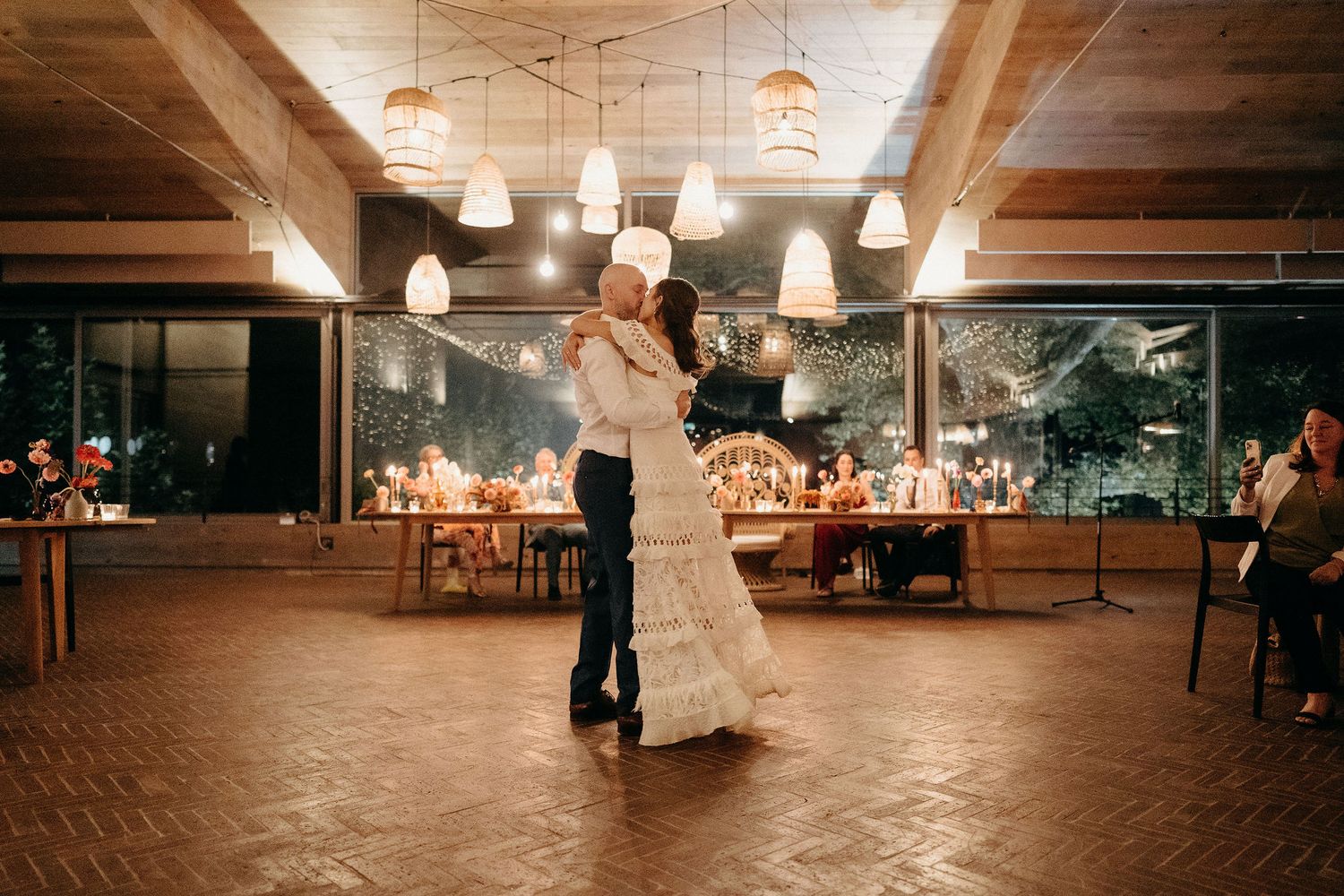 A couple shares their first dance at a wedding reception under warm pendant lights in a rustic venue with wood beams.