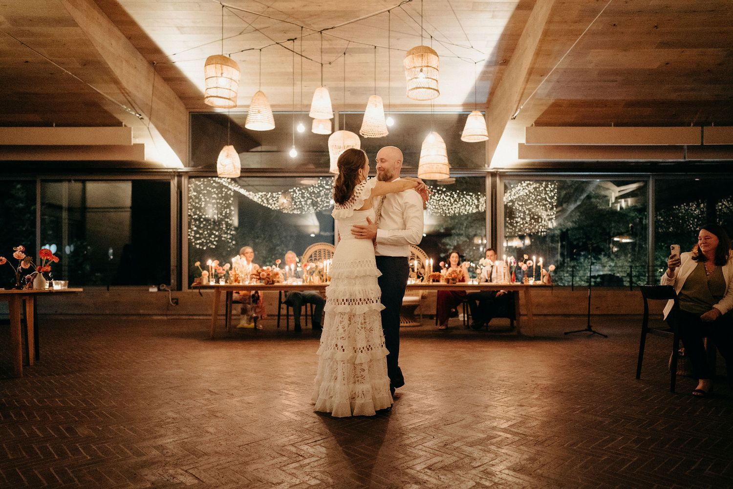 A couple shares their first dance at a rustic indoor wedding reception with pendant lighting and wooden beams.
