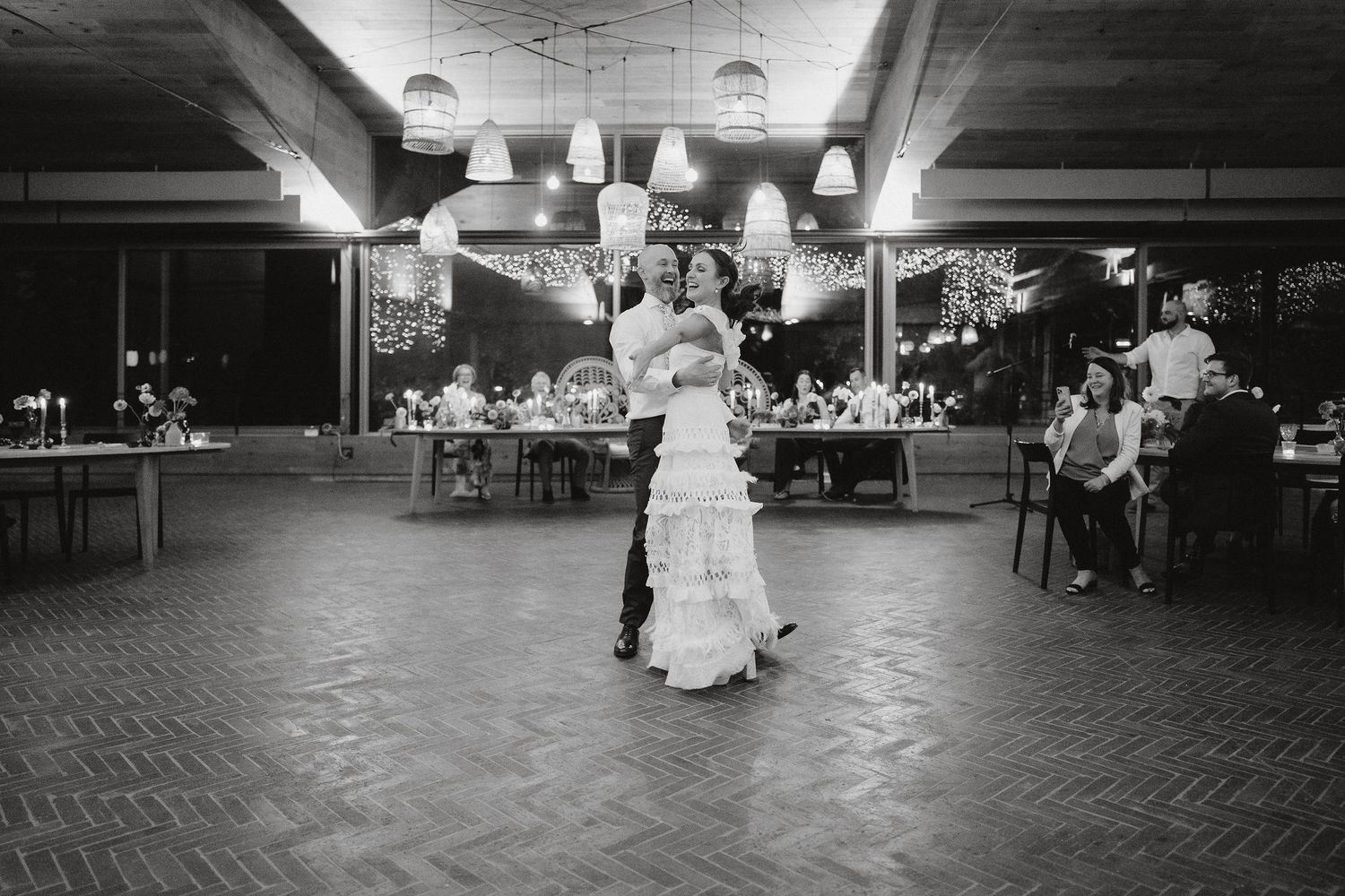 A couple shares a romantic dance at their wedding reception on a brick floor with reception tables visible in background.