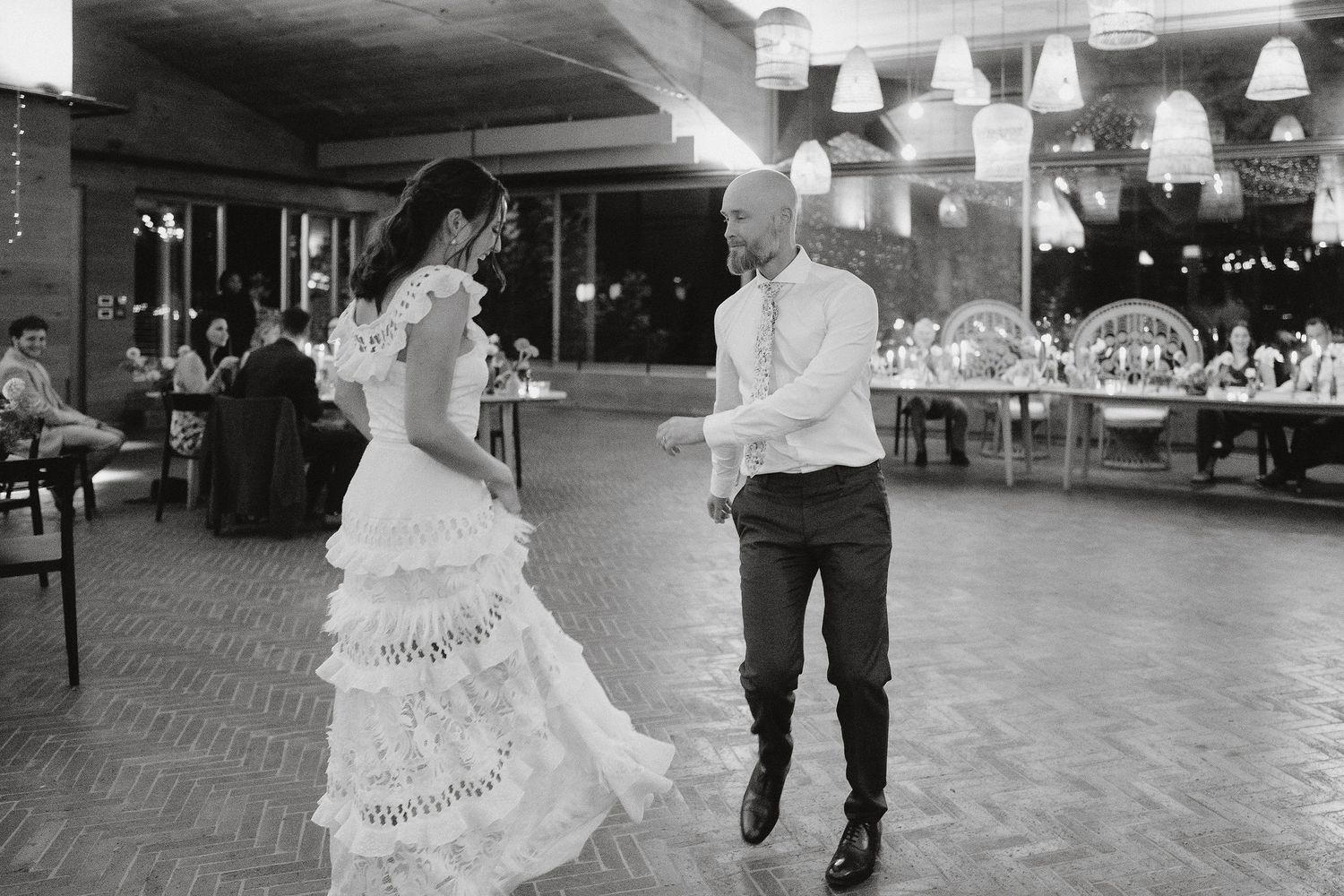 A couple shares their first dance on a reception venue dance floor with hanging pendant lights and tables in the background.
