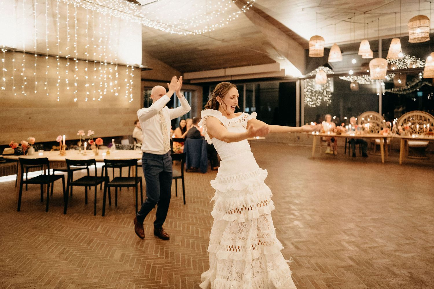 Newlyweds dancing together in a reception hall decorated with string lights and elegant table settings.