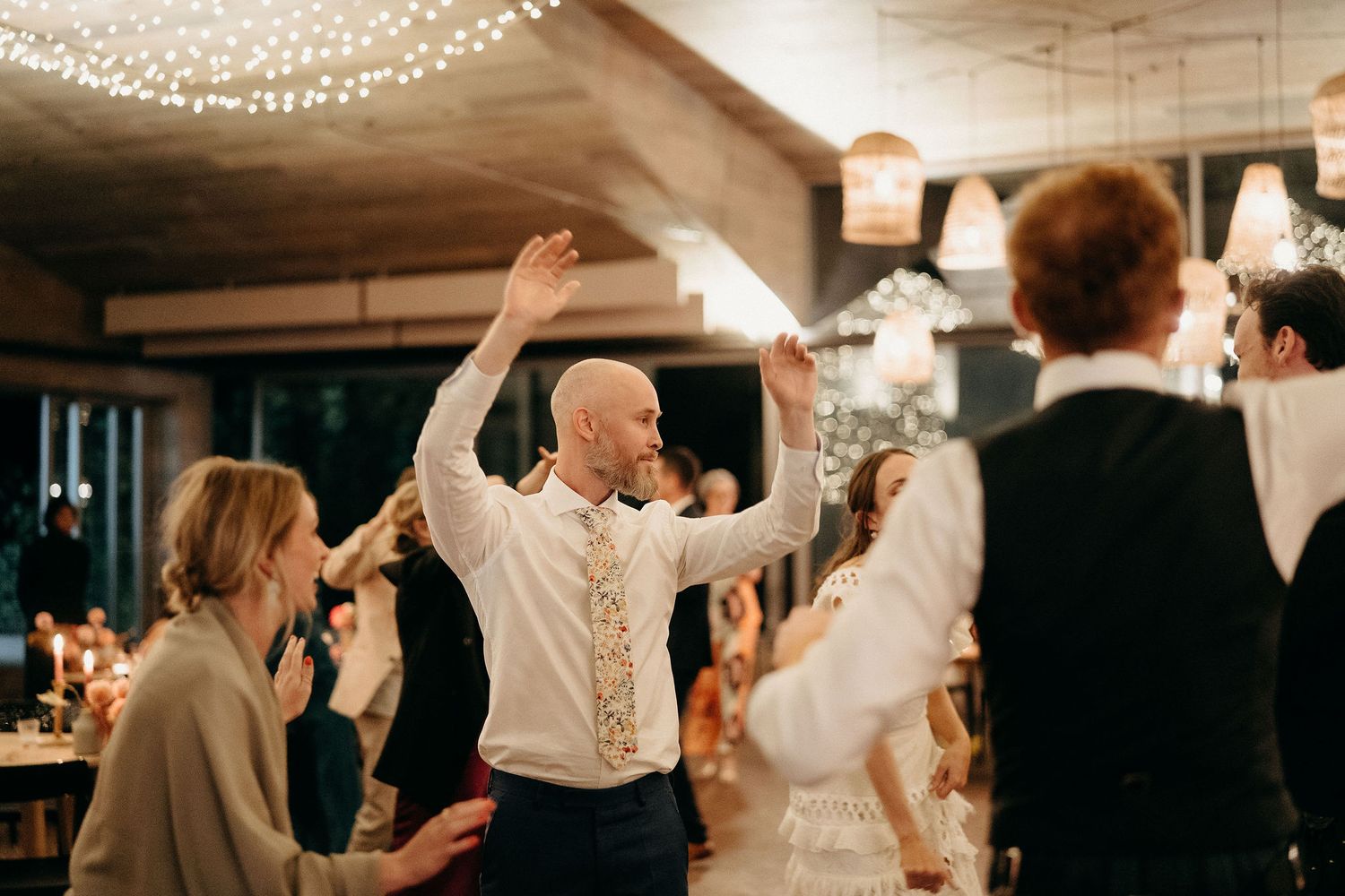 Wedding guests dance and celebrate on the dance floor under string lights at an indoor reception venue.