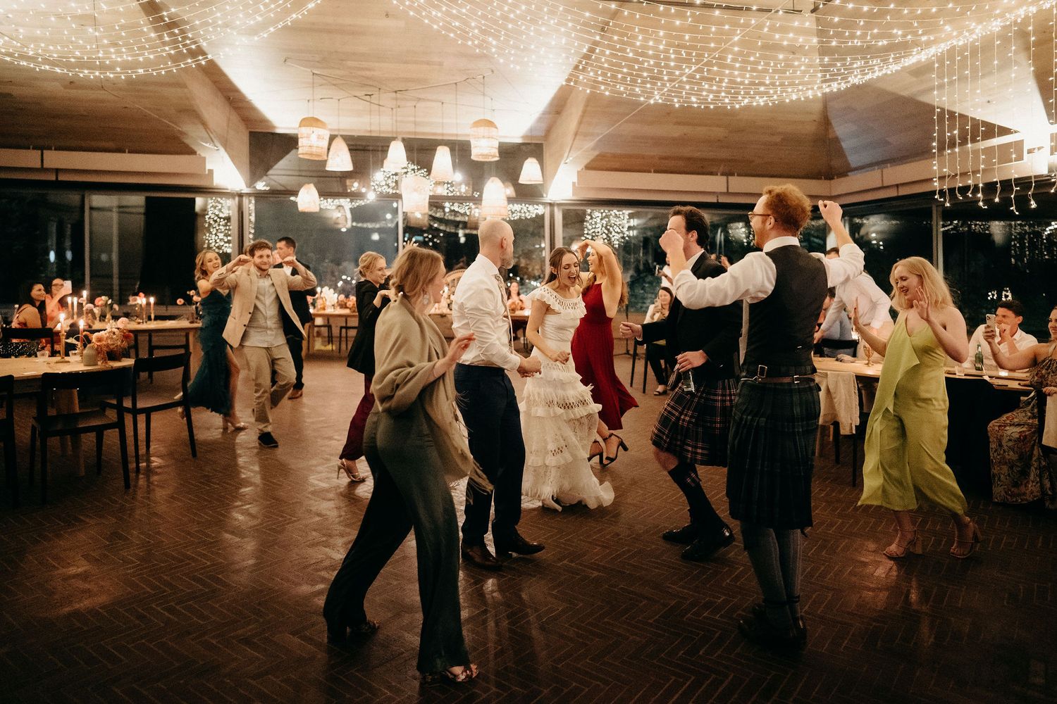 People dance together on a wooden dance floor at an evening celebration with string lights overhead.