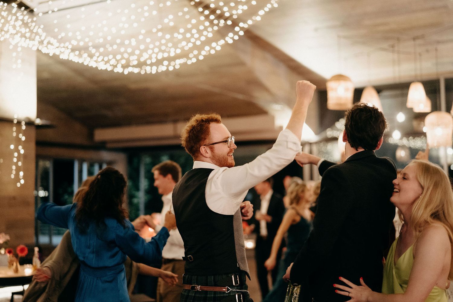 People dancing and celebrating at an indoor event with string lights decorating the rustic ceiling.