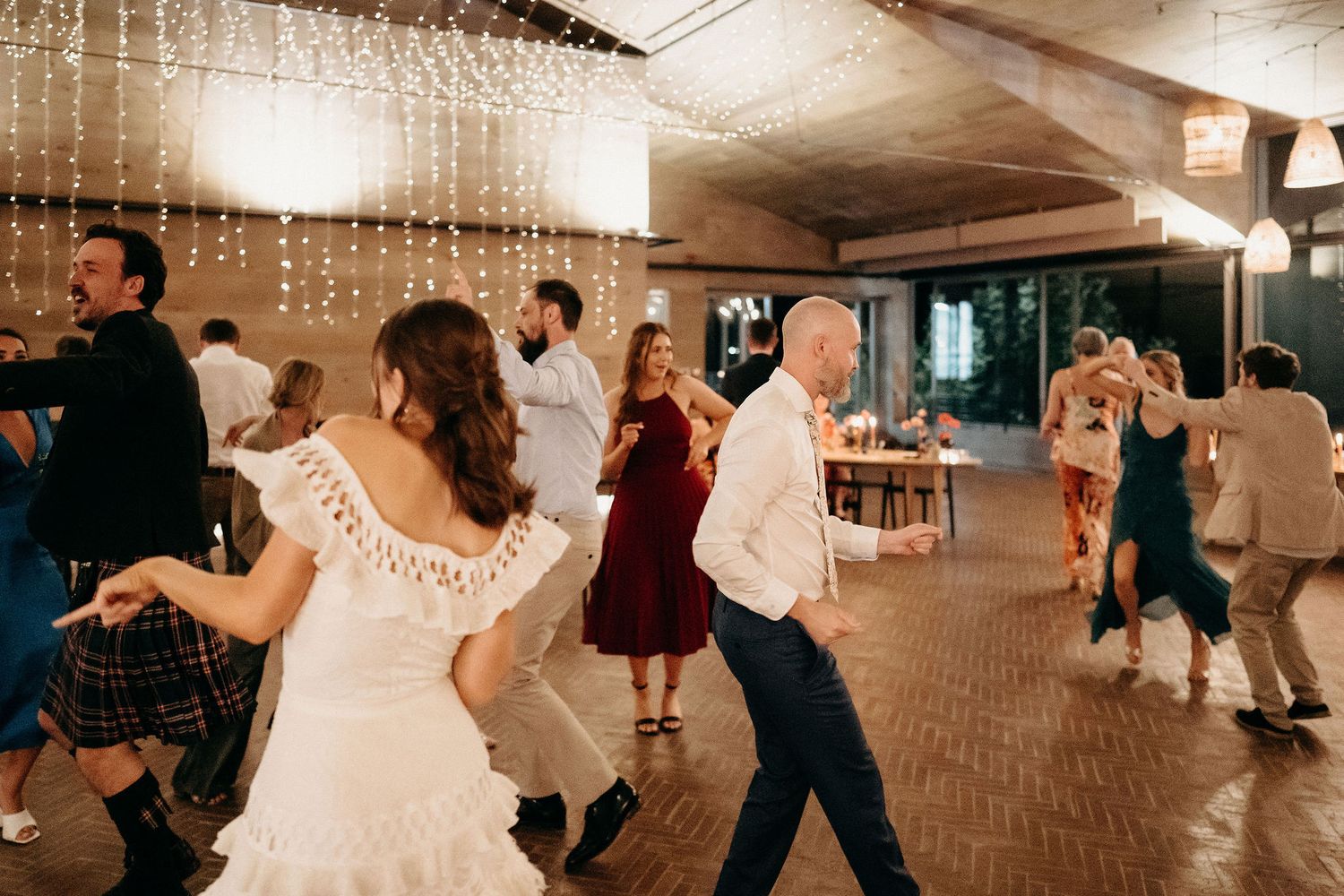 Wedding party dancing energetically during reception celebration with hanging lights in warm tones.