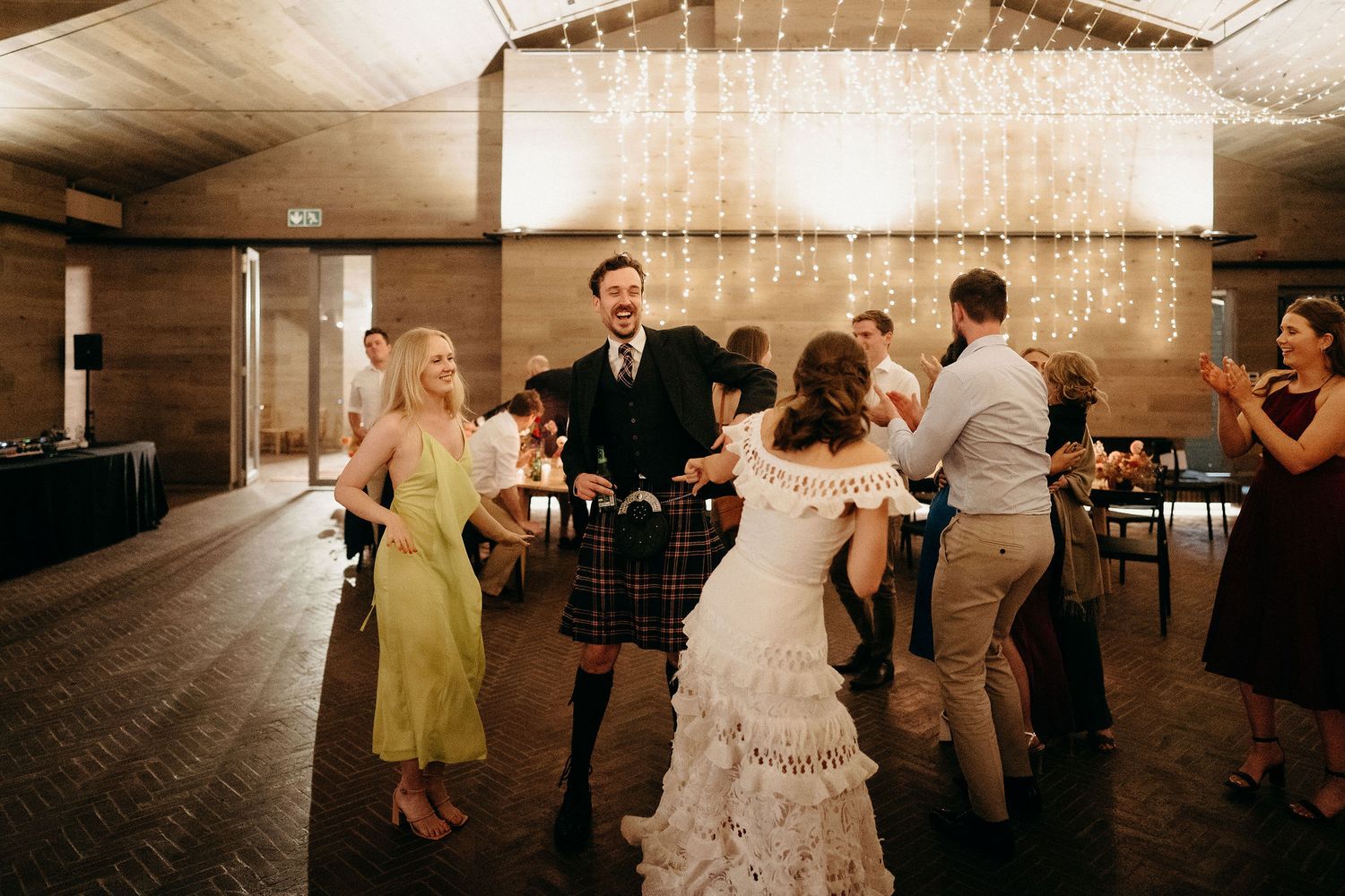Guests dance and celebrate at a rustic indoor wedding reception with string lights illuminating the wooden venue.