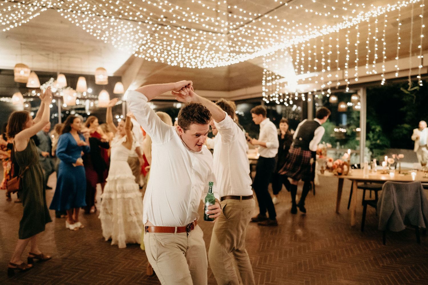 Wedding reception guests dance and celebrate under string lights at an elegant outdoor covered venue.