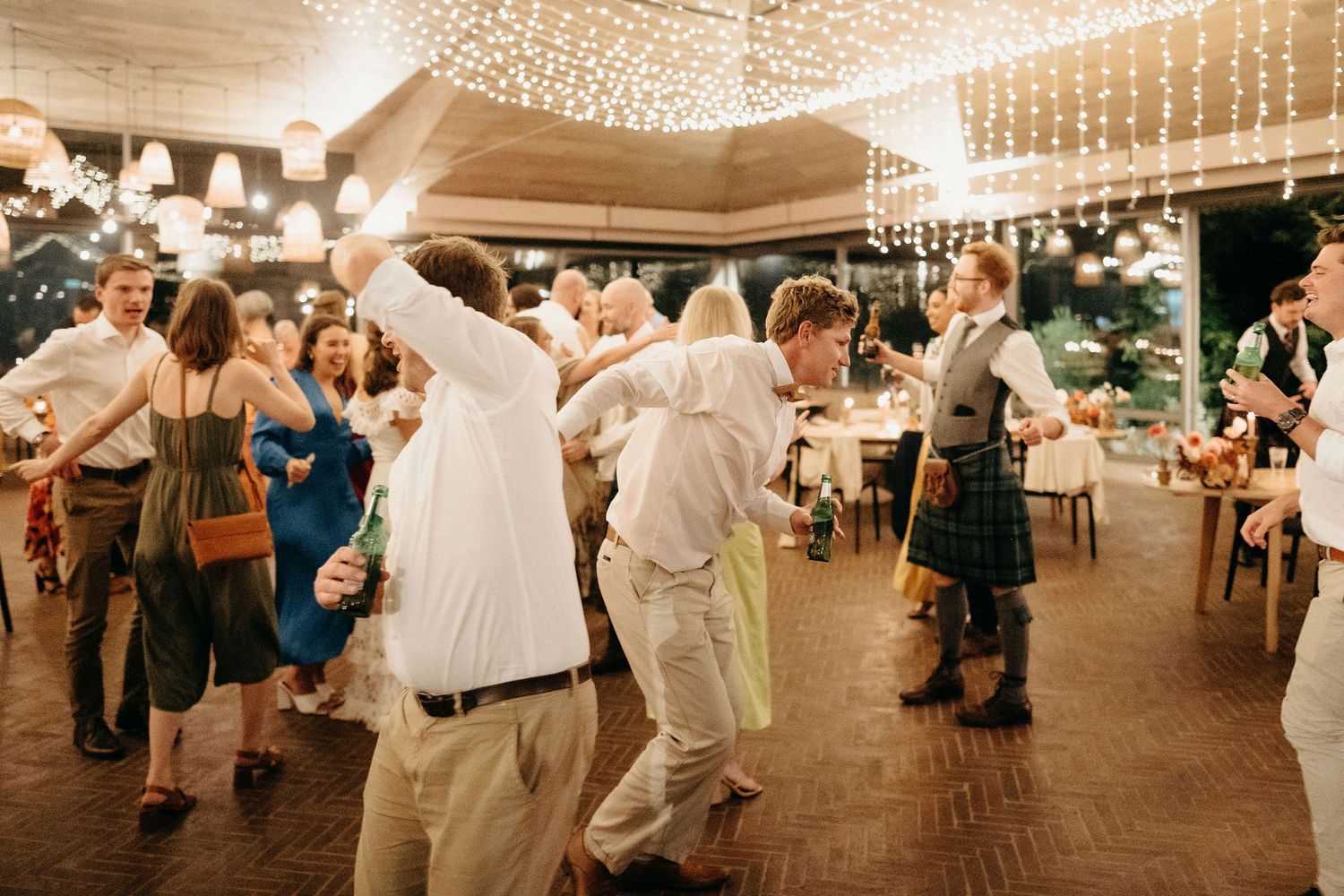 Wedding guests dance together on a wooden dance floor under glowing string lights at a reception venue.