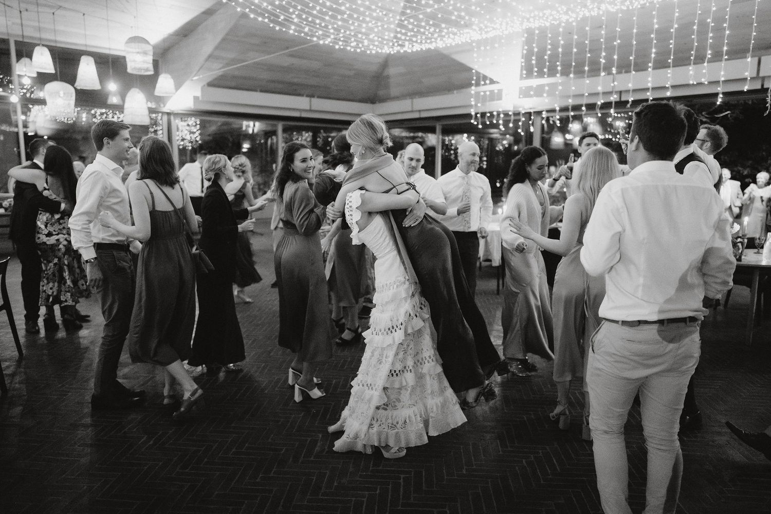 Wedding guests dance together on the dance floor under twinkling lights at an indoor reception venue.
