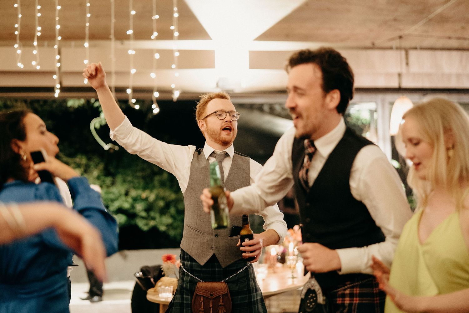 Wedding guests celebrate joyfully at an outdoor reception with string lights overhead and a person in a kilt raising their arm.