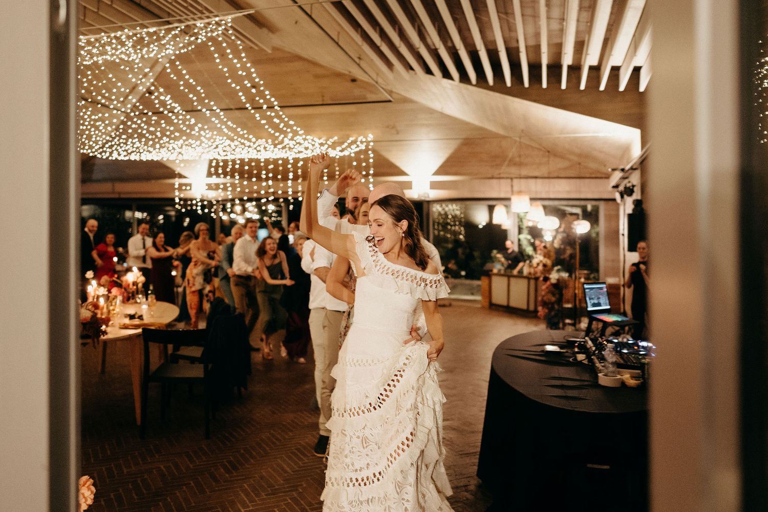 A couple shares a dance moment at their wedding reception under decorative string lights in a warm-lit venue.