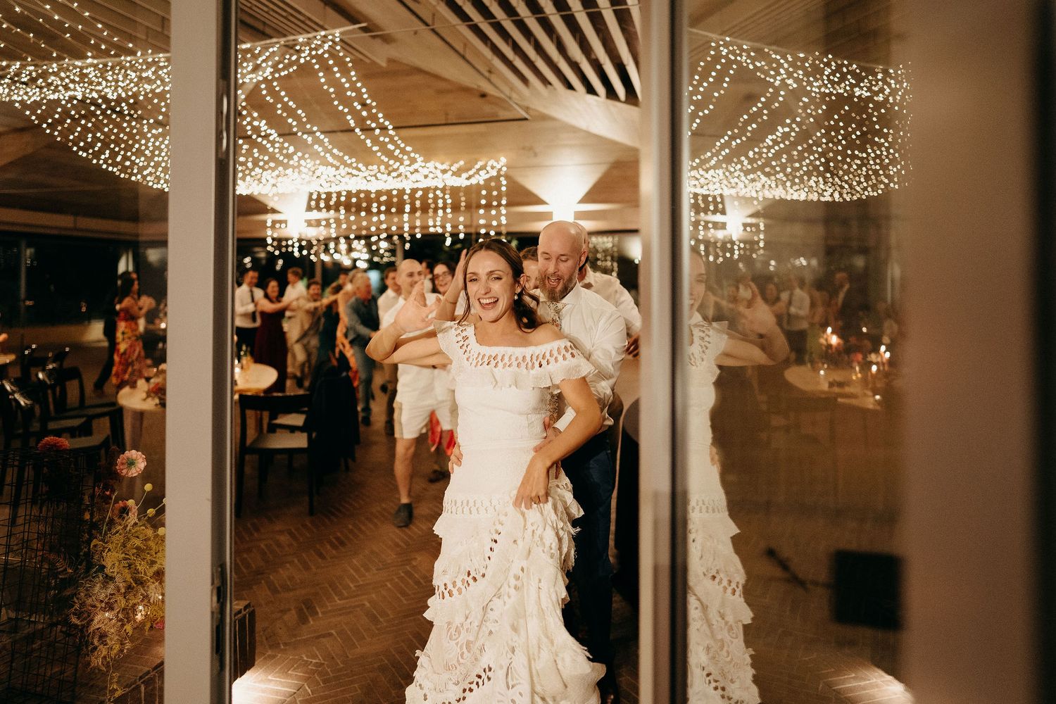 A lively indoor reception with twinkling string lights overhead illuminates dancers through glass windows at night.