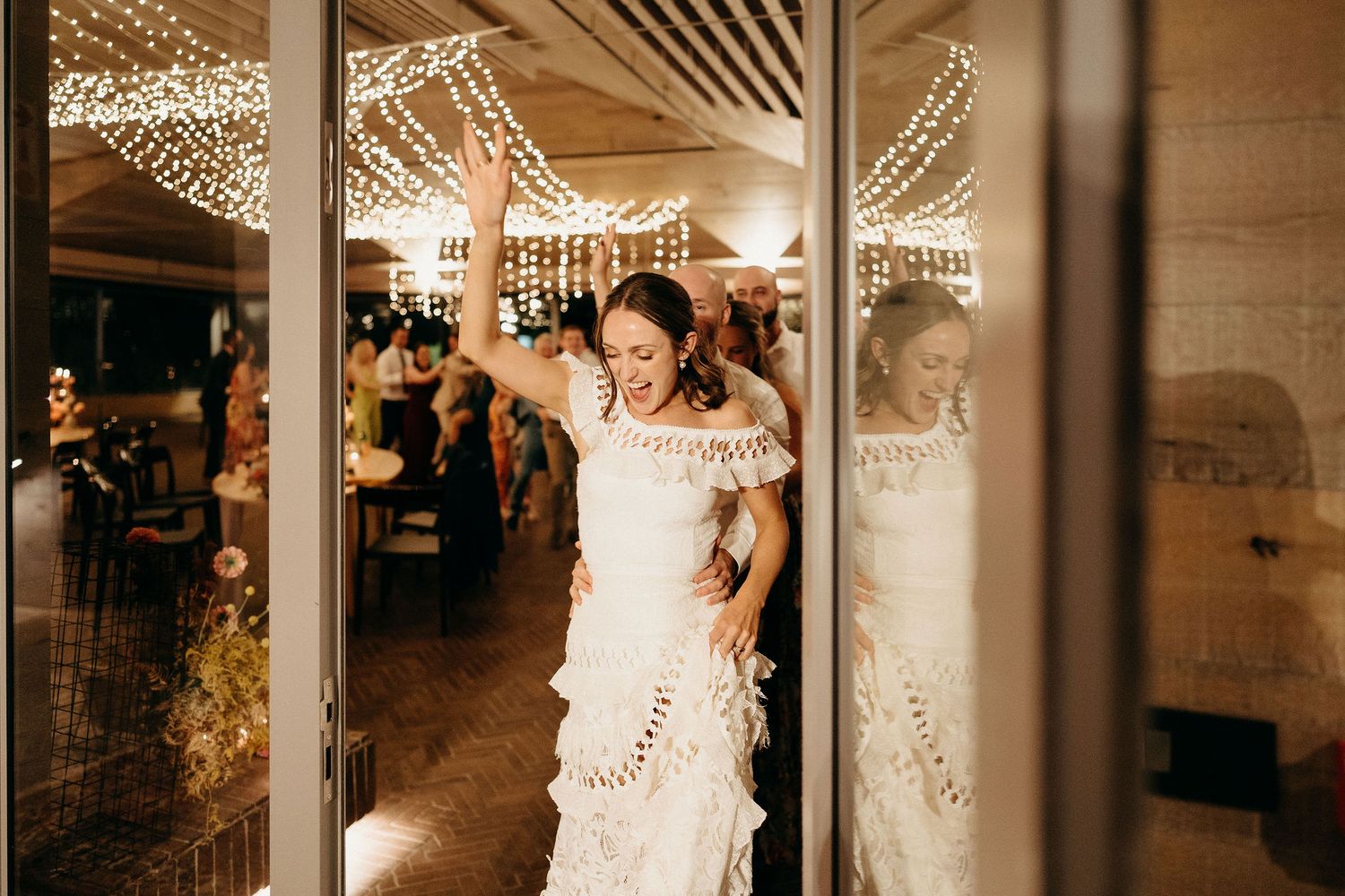 A bride in a lace wedding dress stands in a reflective glass doorway under warm twinkle lights at an indoor reception.