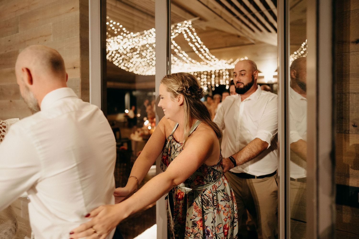 Wedding guests dance and celebrate on a dance floor illuminated by warm string lights in an indoor venue.