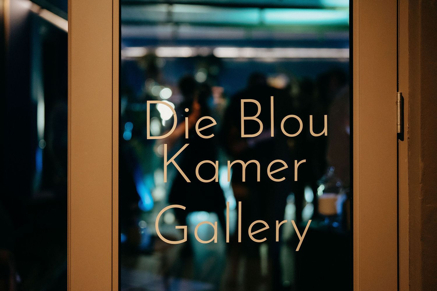 Gold lettering spelling 'Die Blou Kamer Gallery' on a glass window with blue lighting behind.