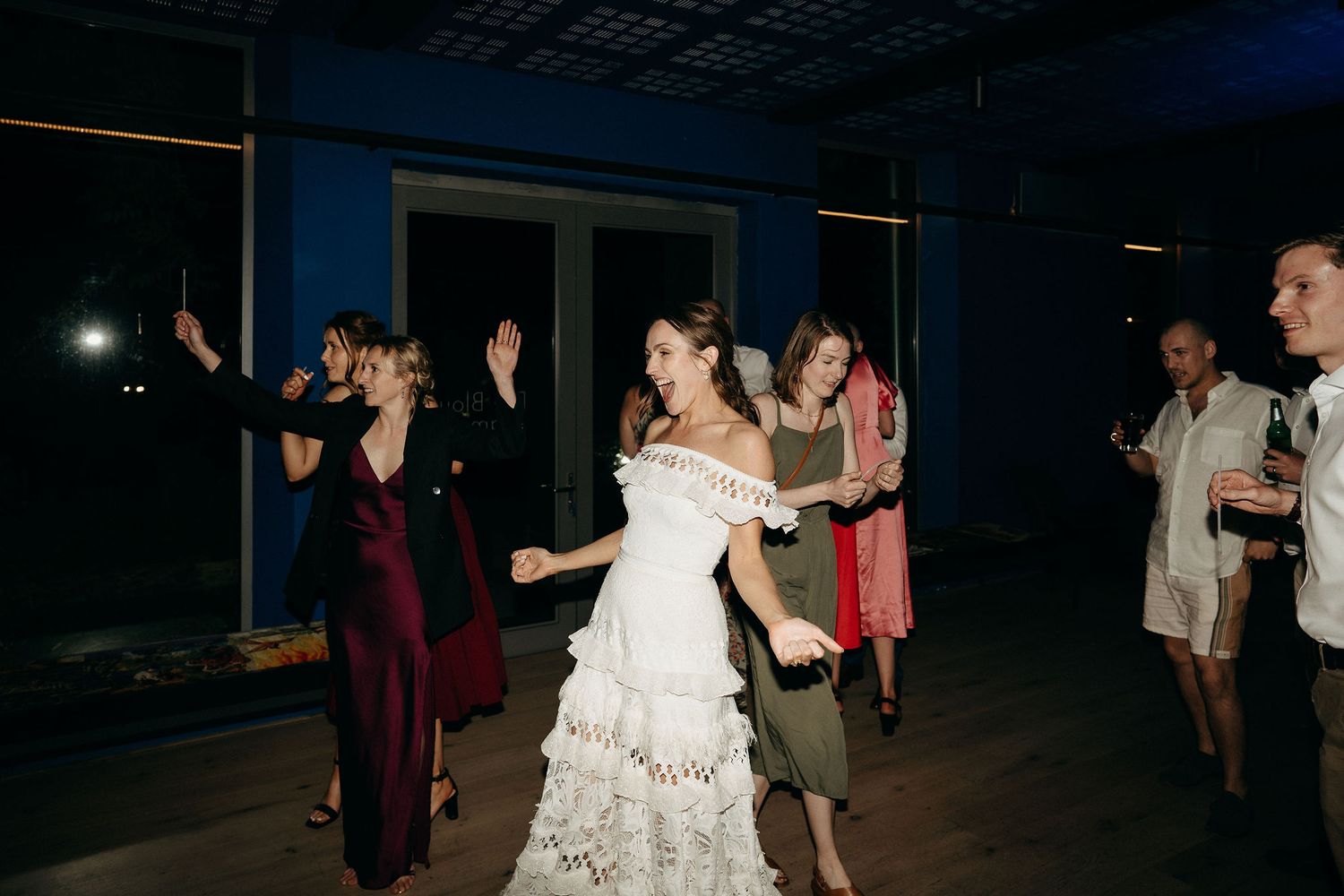 Wedding party dances and celebrates during nighttime reception on dark dance floor with decorative lighting.