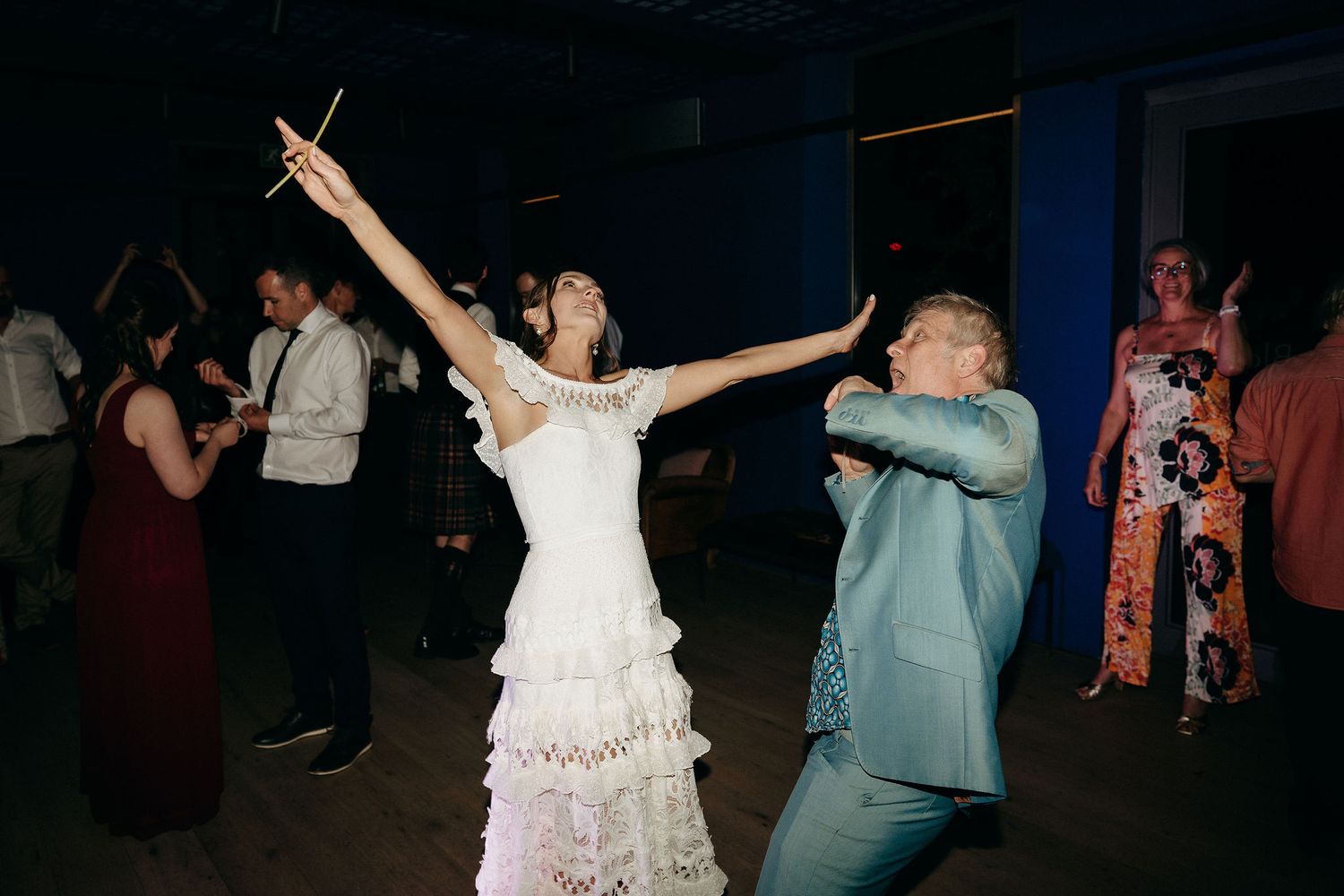 Wedding guests dancing enthusiastically on a dark dance floor with the bride in a white ruffled dress and arms raised.