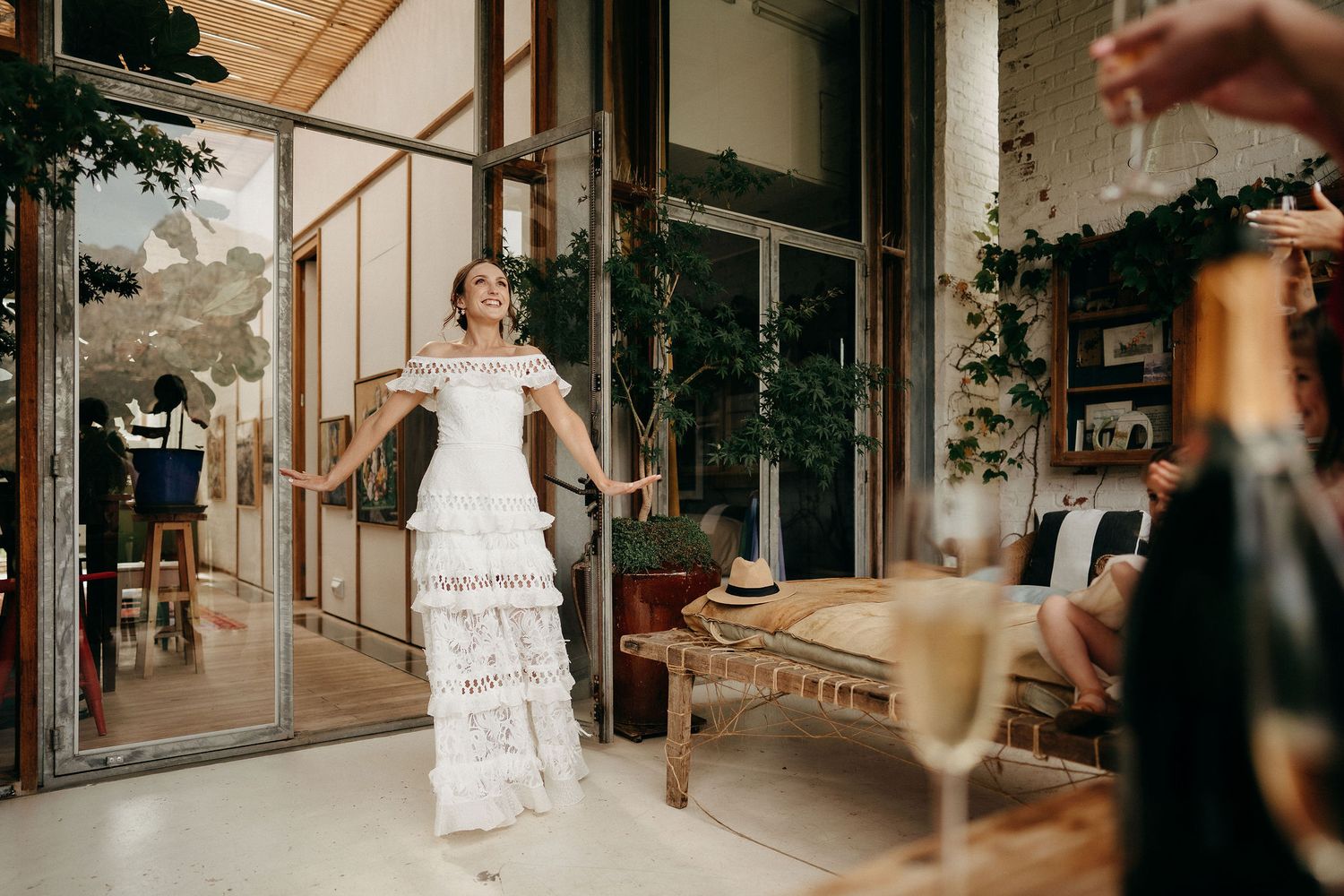 A bride in a layered white wedding dress with ruffles stands in front of a rustic venue entrance with greenery.